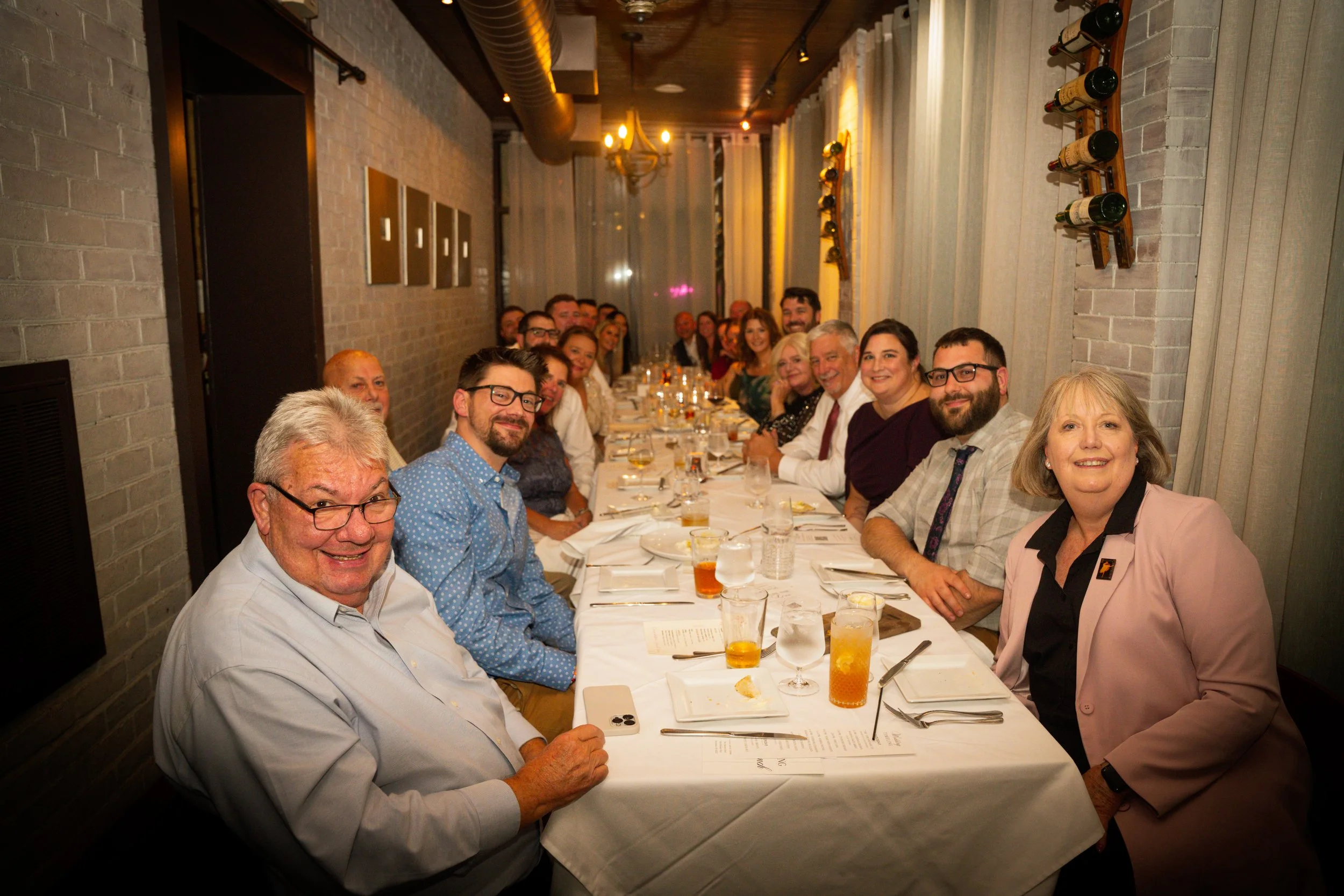 A long dinner table with about thirty people gathered for a meal in a restaurant with brick walls and white curtains. The guests are smiling and looking toward the camera, with glasses of drinks and menus on the table.
