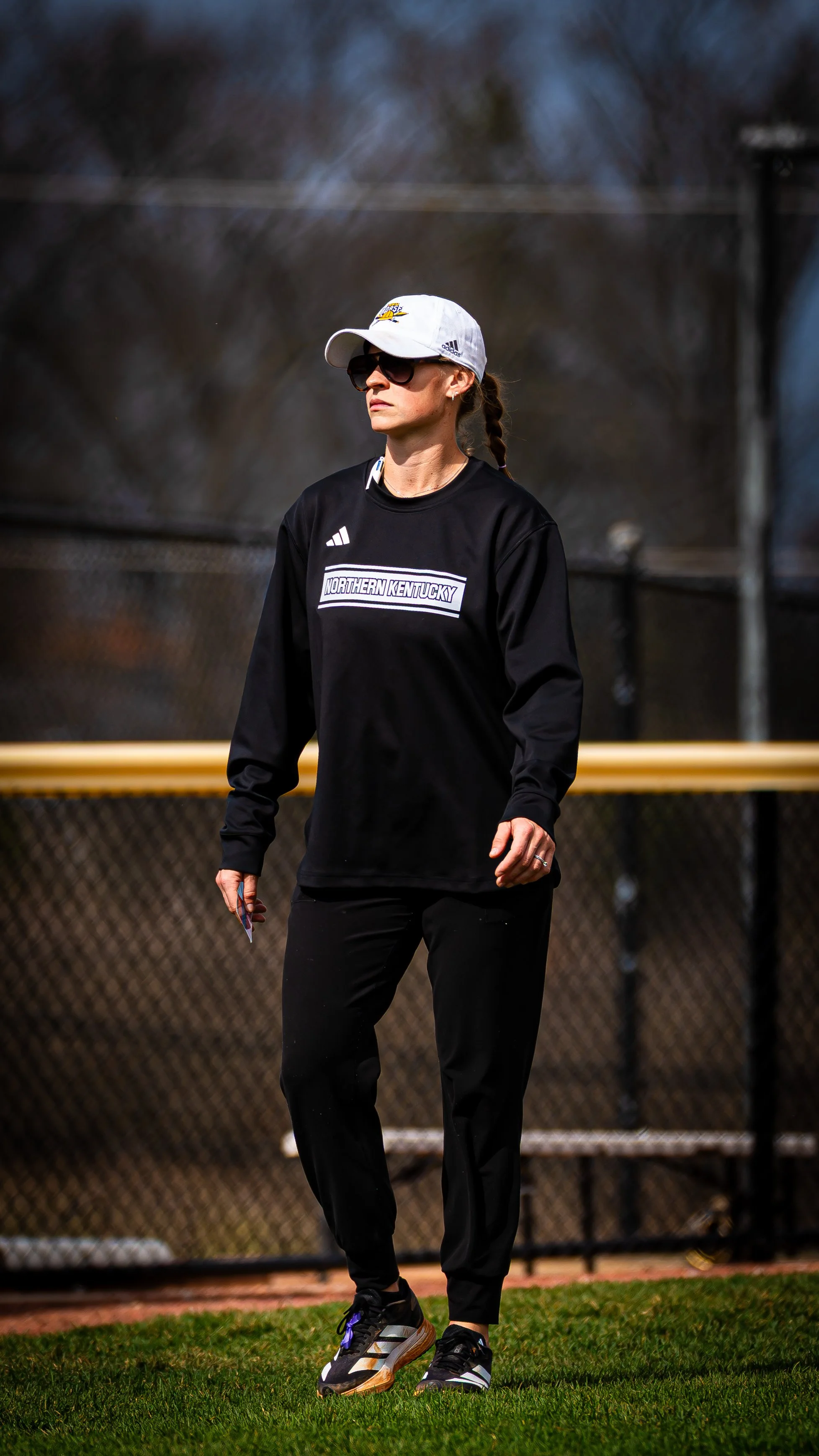 A woman wearing athletic clothing, sunglasses, and a white cap stands on a baseball field with a chain-link fence in the background.