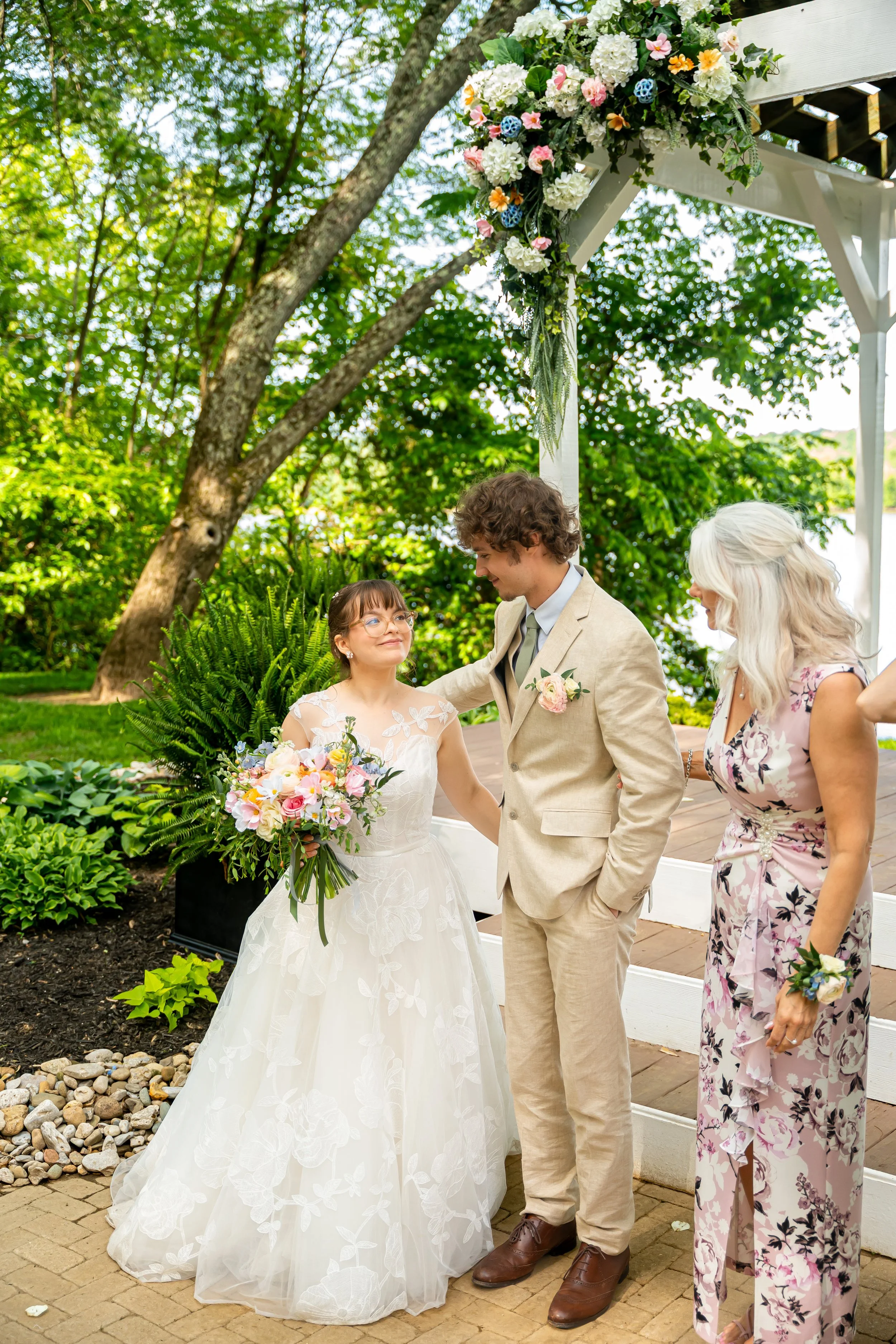 A wedding ceremony outdoors with a bride, groom, and two women, under a white arch decorated with flowers, lush green trees, and a natural setting.