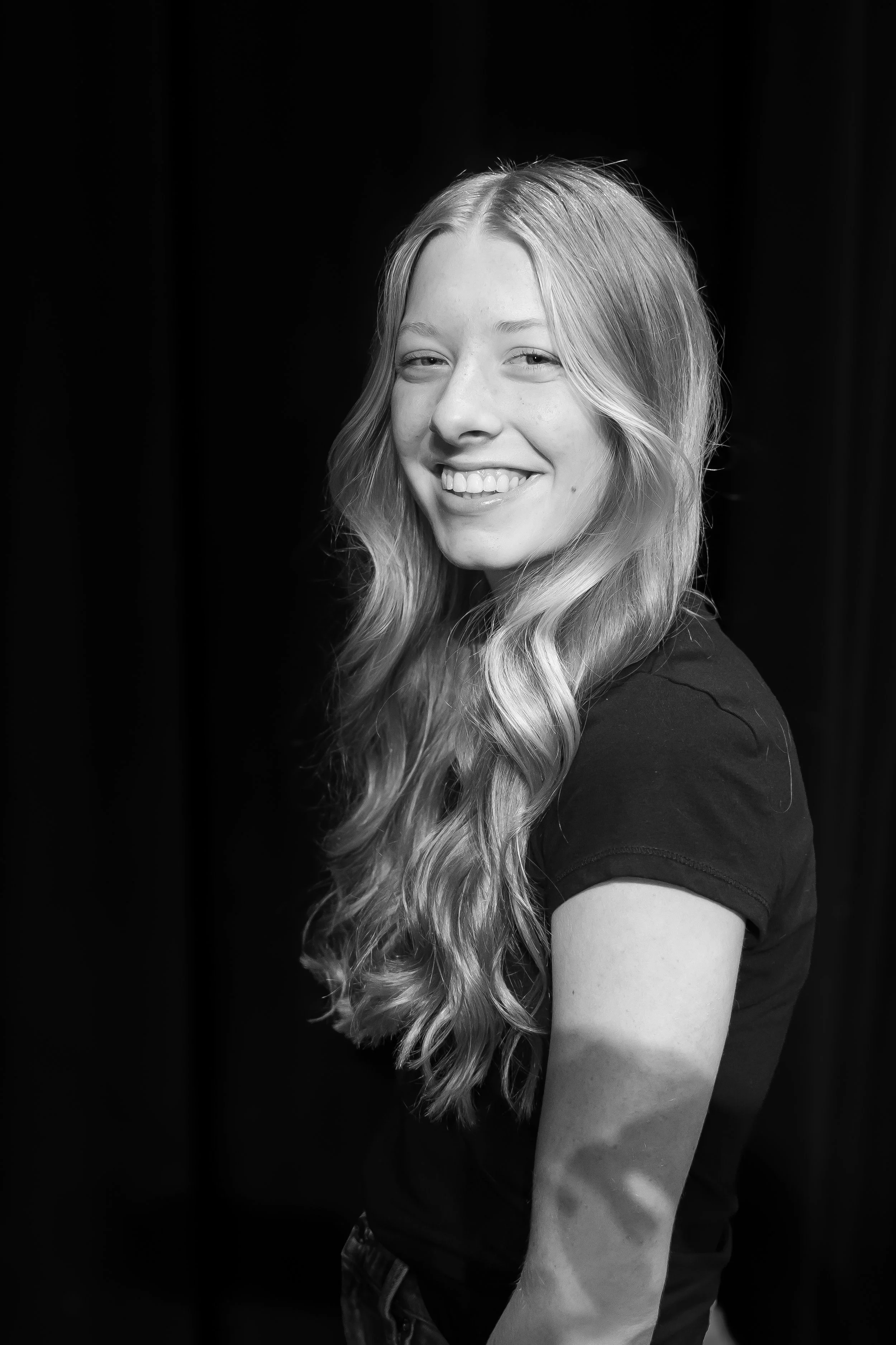 Black and white portrait of a young woman with long wavy hair, smiling, against a dark background.