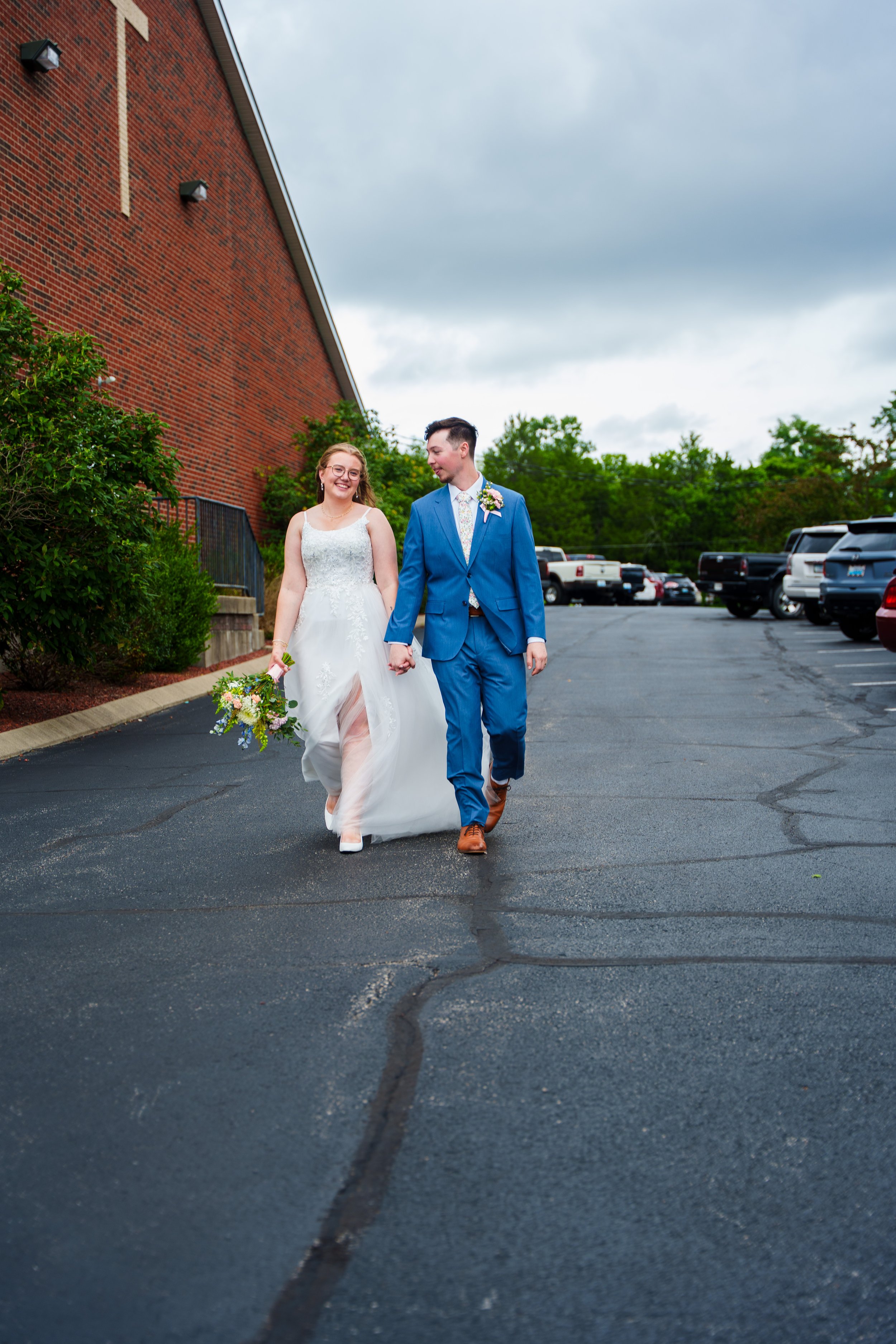 A newlywed couple walking hand in hand on an outdoor asphalt parking lot. The bride is dressed in a white wedding gown holding a bouquet, and the groom is in a blue suit with a floral tie and boutonnière. The parking lot is lined with parked vehicles