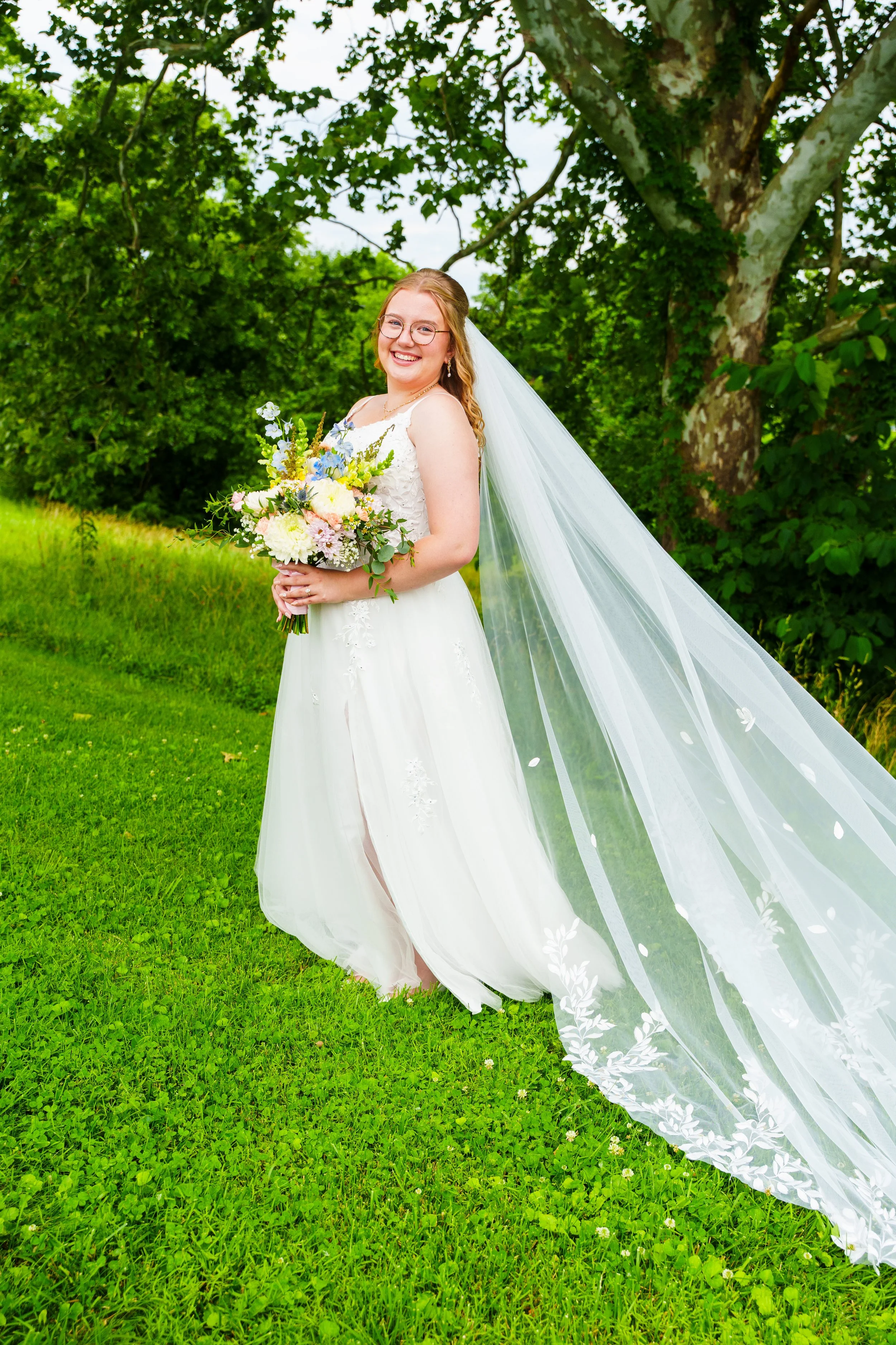 A smiling bride with glasses in a white wedding gown holding a bouquet of flowers, standing outdoors on green grass with trees in the background and her veil flowing behind her.