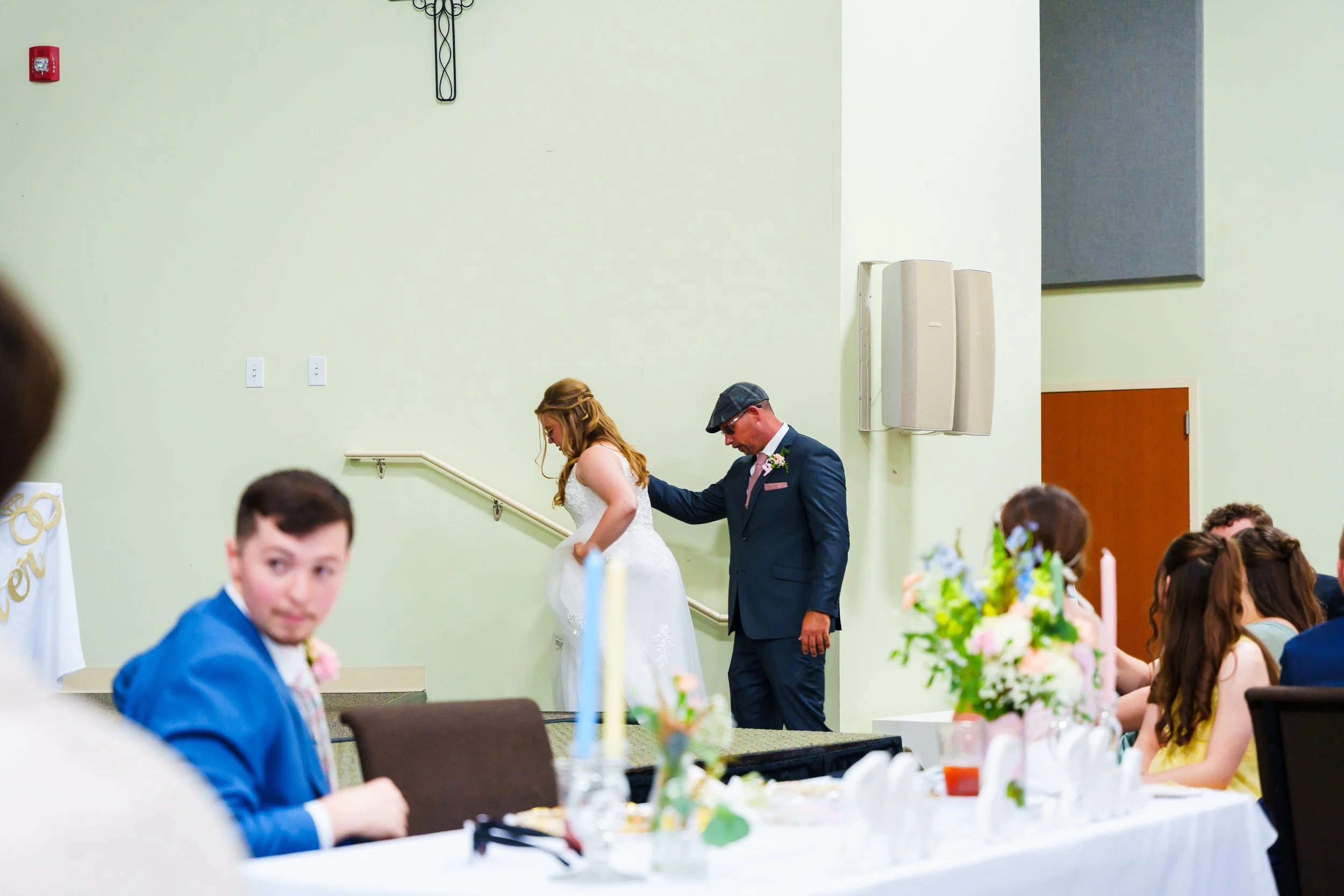 A bride and groom holding hands while walking down the aisle during a wedding ceremony inside a church or hall, with wedding guests seated at tables decorated with flowers and candles.