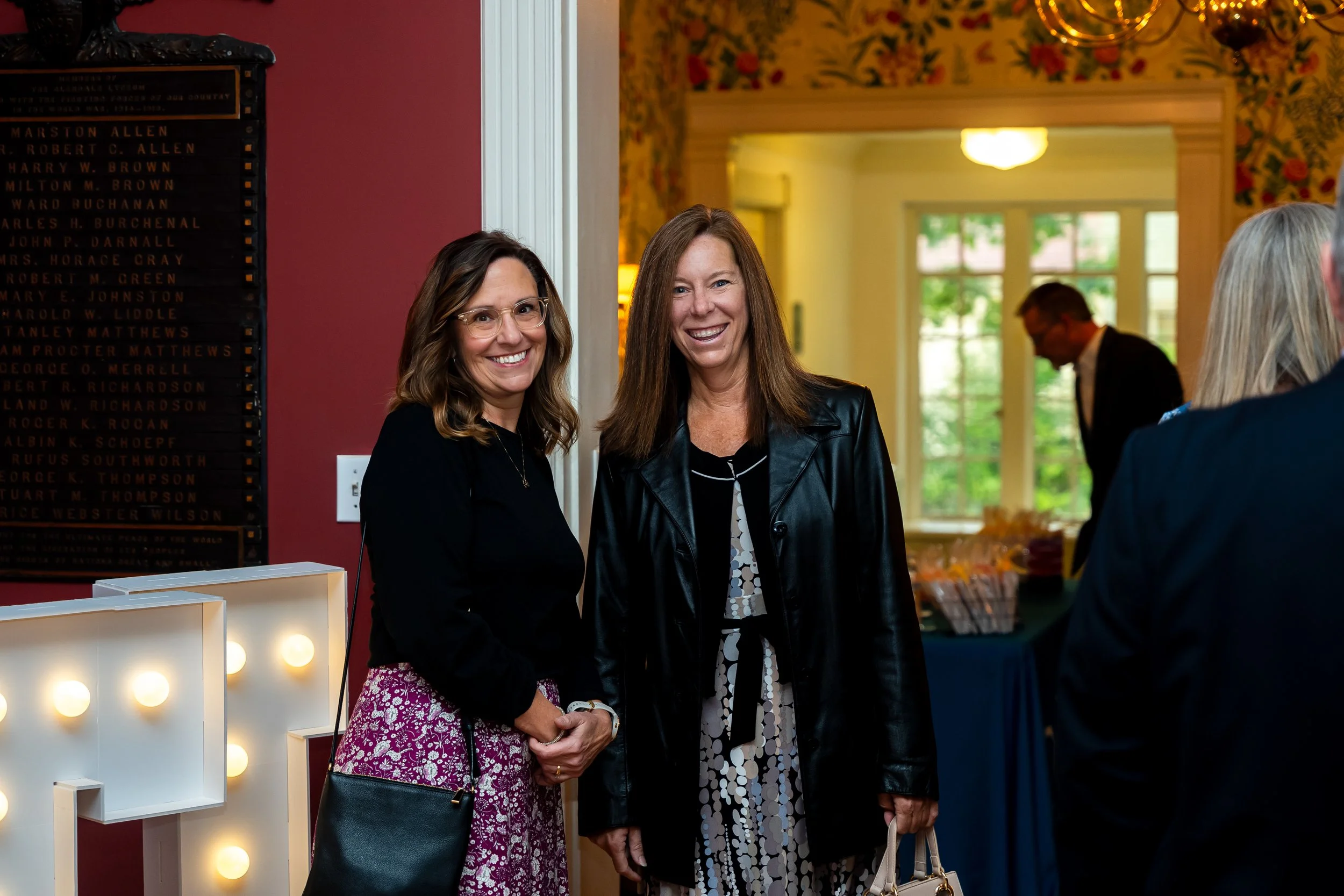 Two women smiling and talking at an indoor event, with one woman wearing glasses and a black top and the other woman wearing a black leather jacket and a patterned dress, while people are visible in the background.
