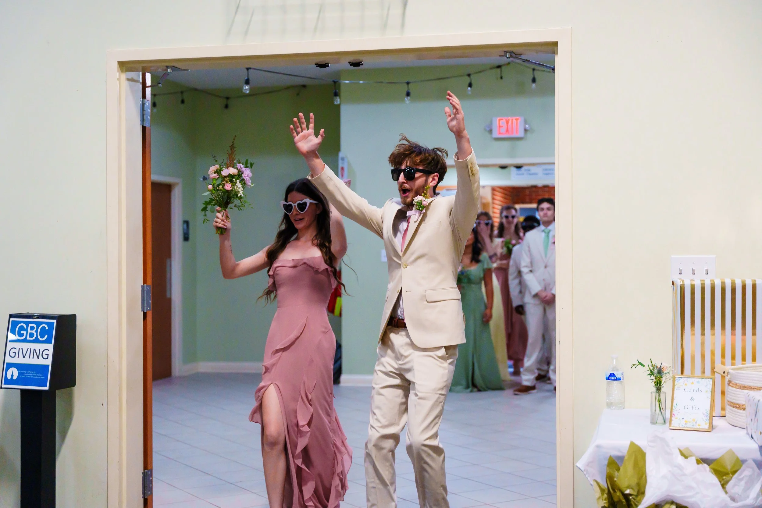 A wedding celebration with a stylish couple walking through an entrance. The woman wears a pink dress and heart-shaped sunglasses, carrying a bouquet. The man wears a beige suit, sunglasses, and is raising his hands. In the background, guests are dre