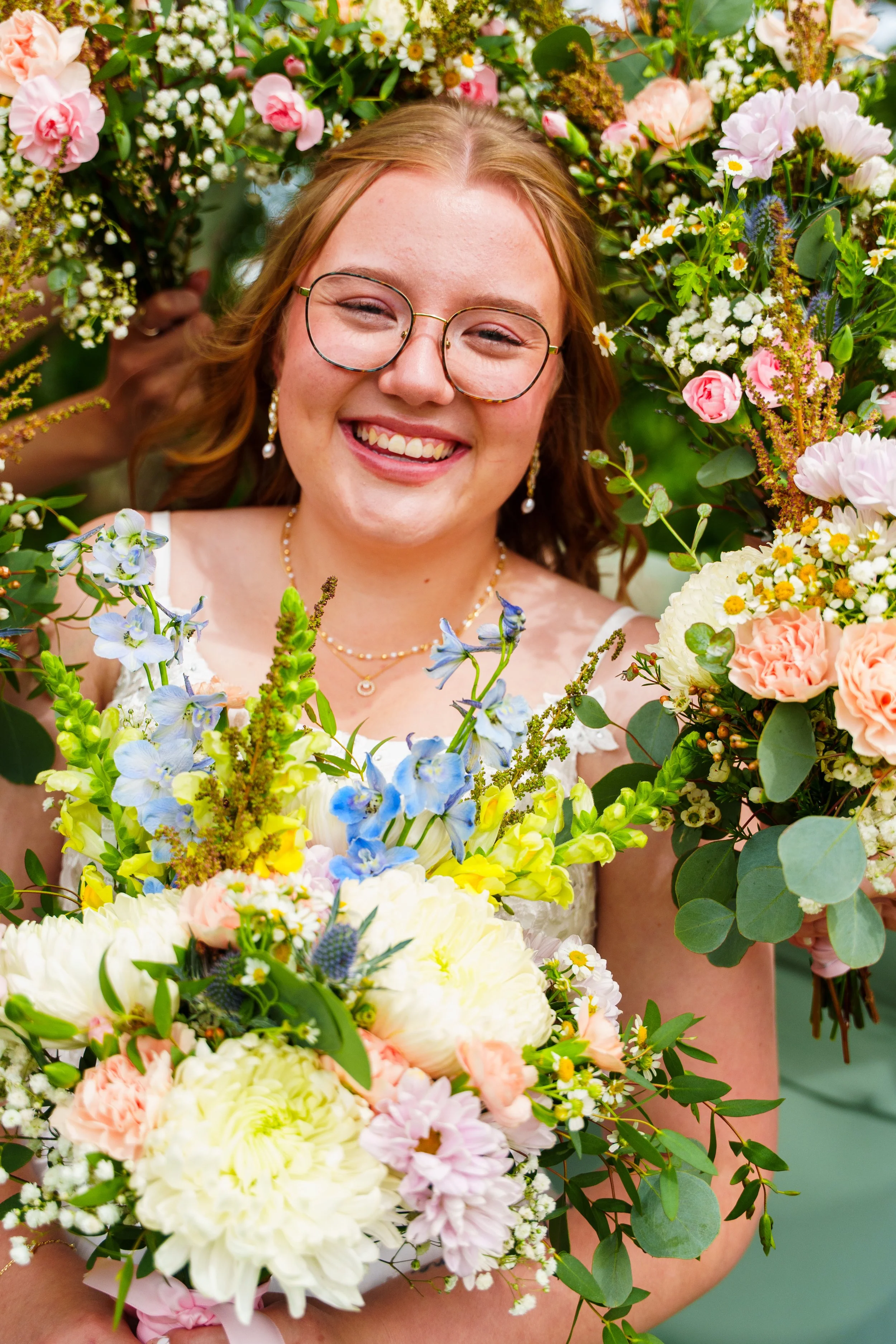 A woman with glasses smiling, surrounded by a large bouquet of colorful flowers including peonies, roses, and daisies.