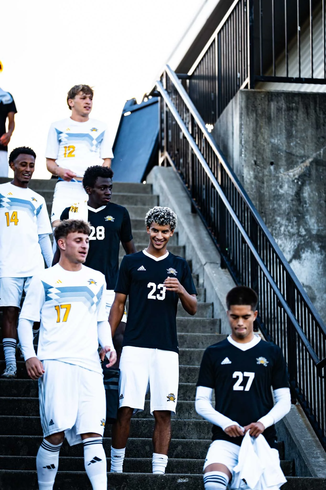 Group of young male soccer players descending concrete stairs outdoors, wearing athletic uniforms, some in black shirts and others in white, with a black fence on the right side of the image.