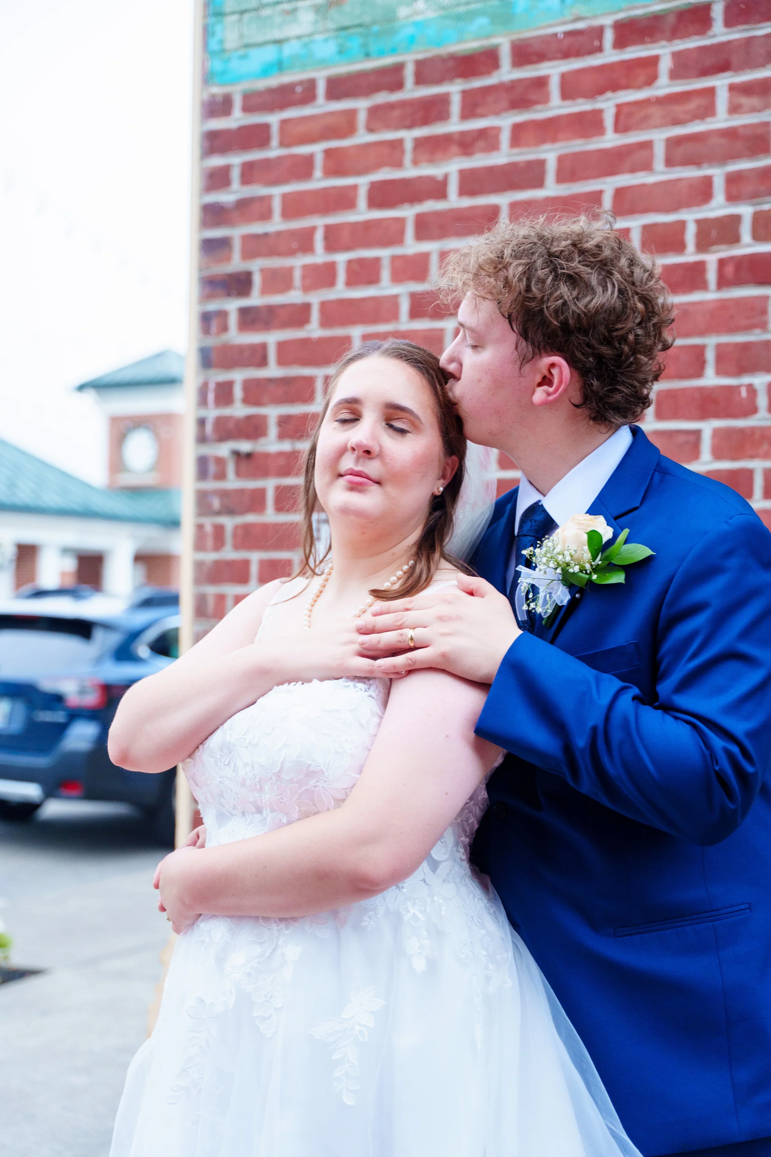 A bride and groom sharing a dance outside on a cloudy day. The groom is kissing the bride on the forehead while she has her eyes closed and arms crossed, wearing a white lace wedding dress and pearl necklace, with a red brick wall behind them.