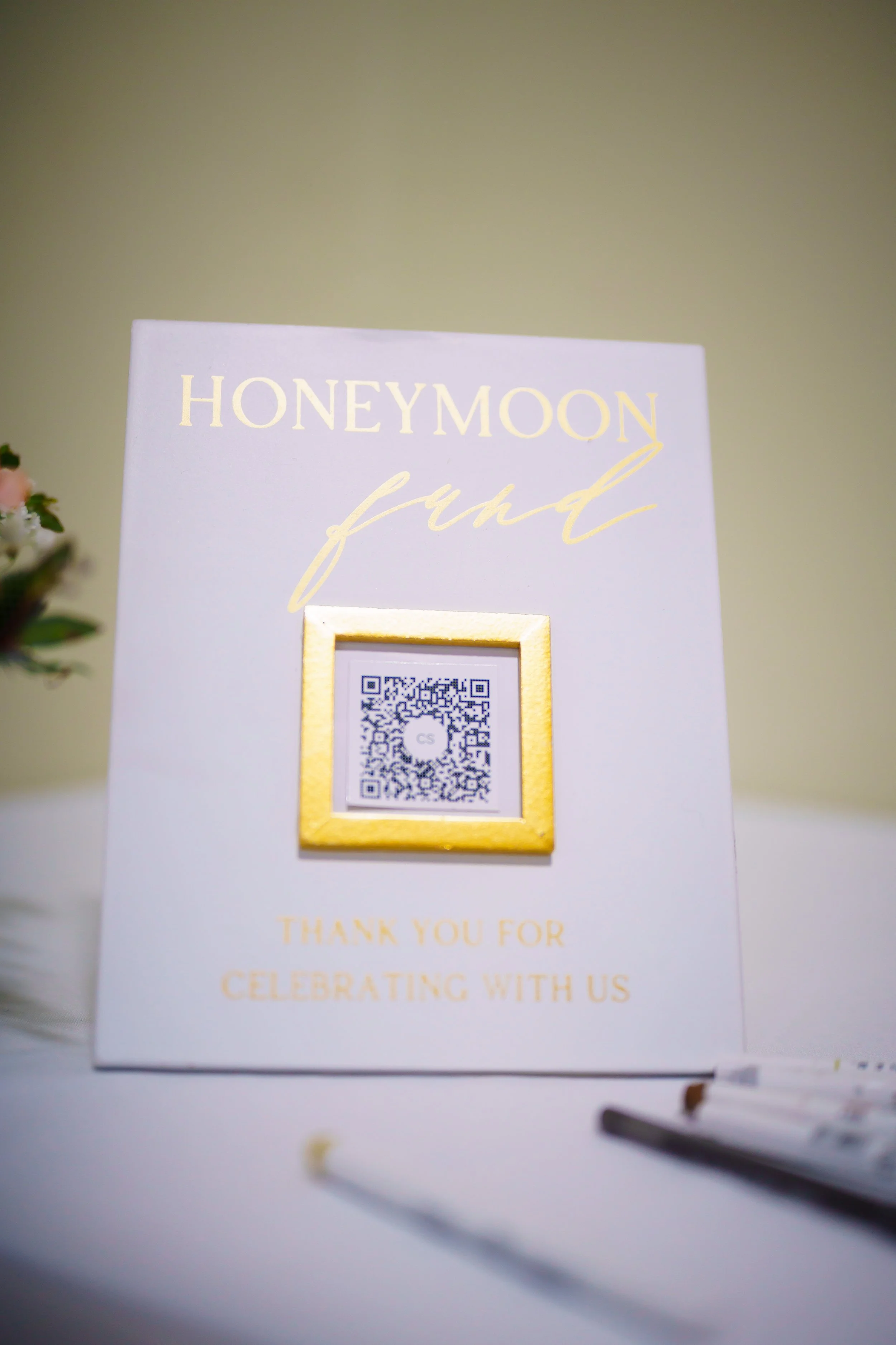 A white card with gold lettering that says 'HONEYMOON fund' and a QR code beneath it, set against a green background with some flowers partially visible on the left side.