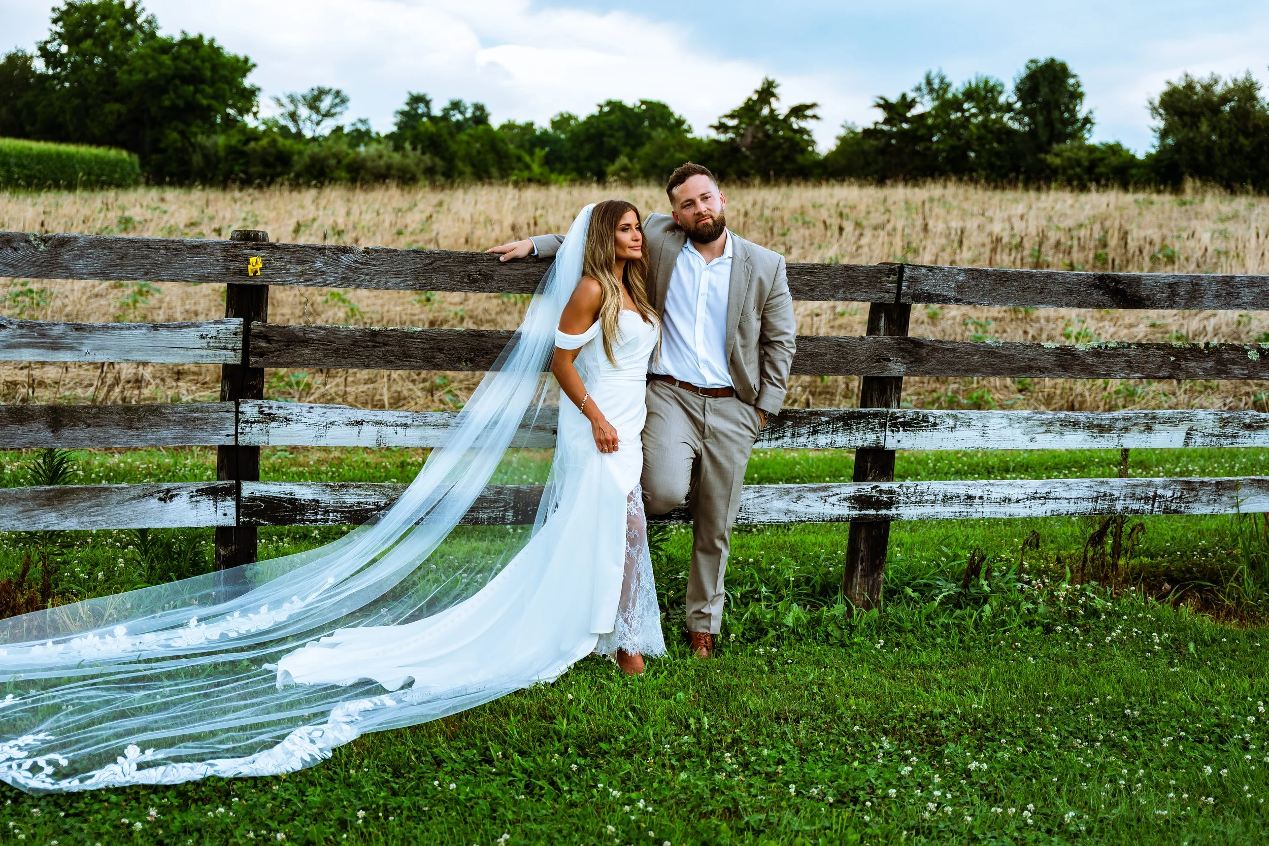A bride and groom posing outdoors near a wooden fence, with a field and trees in the background. The bride is wearing a white wedding dress with a veil, and the groom is wearing a light gray suit with a white shirt. The bride has long, wavy blonde ha