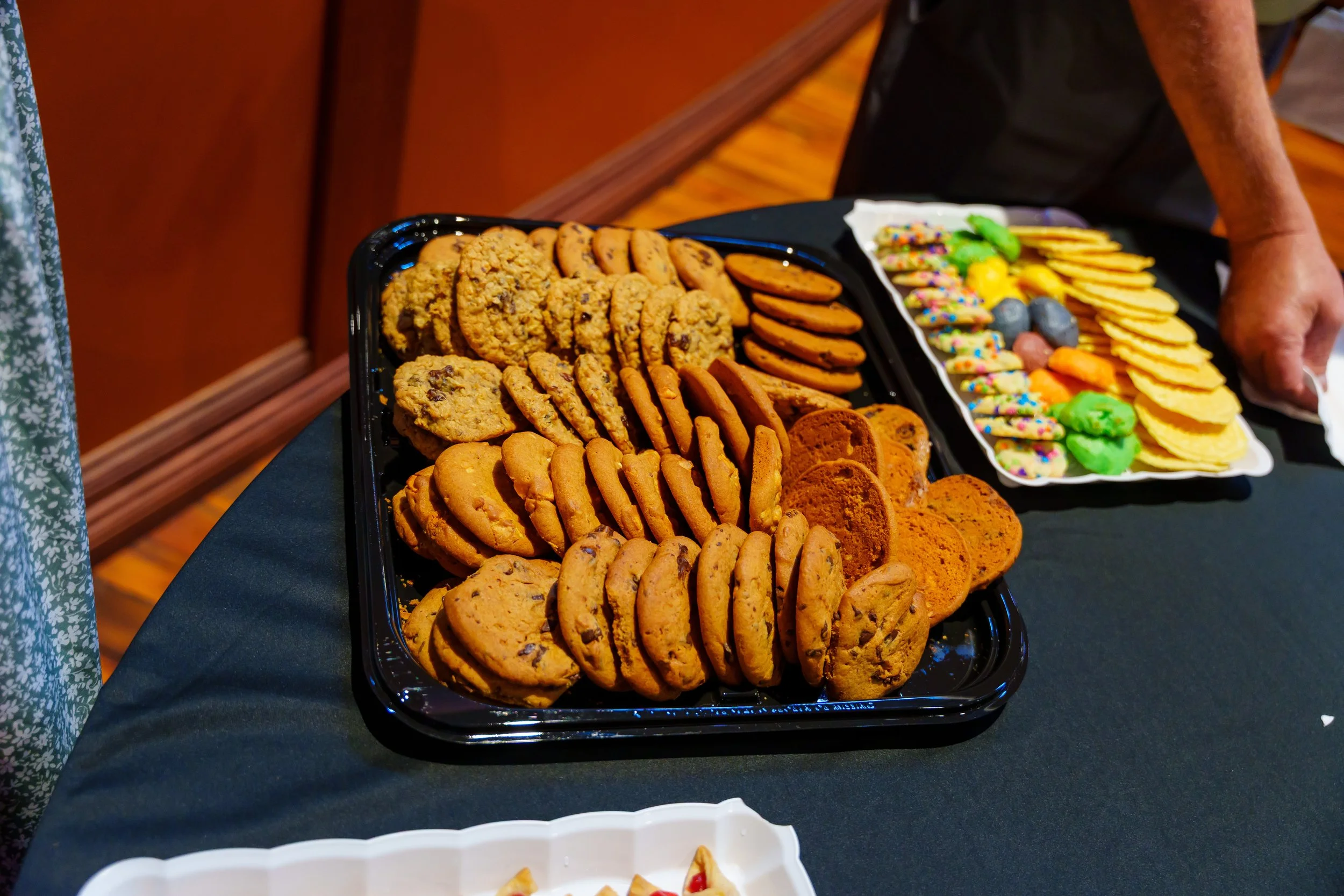 Assorted cookies on a platter, including chocolate chip, oatmeal, and peanut butter cookies, with colorful sprinkles and candy on a separate tray in the background.
