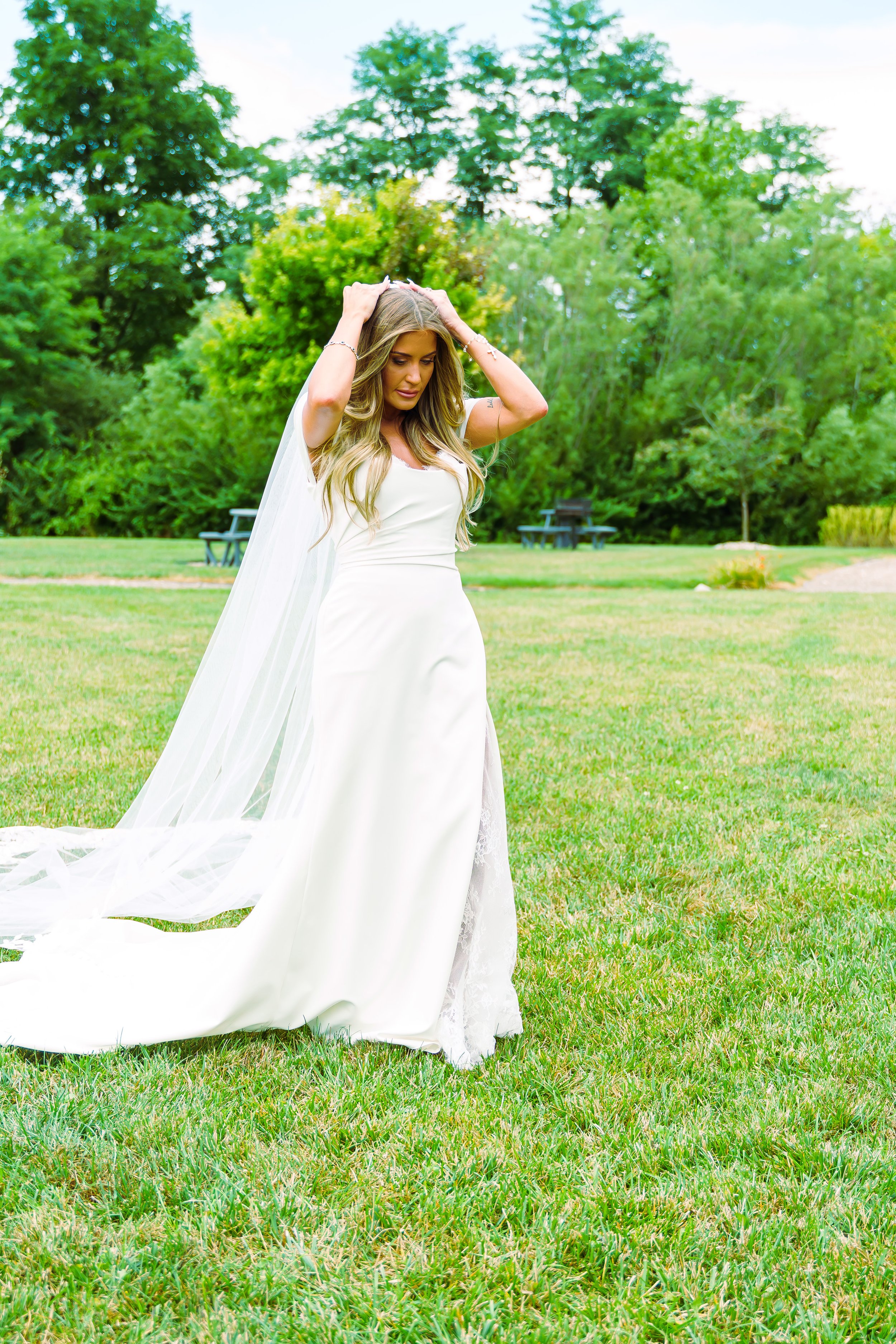 A bride in a white wedding dress standing on a grassy field with her hands on her head, surrounded by green trees, during daytime.