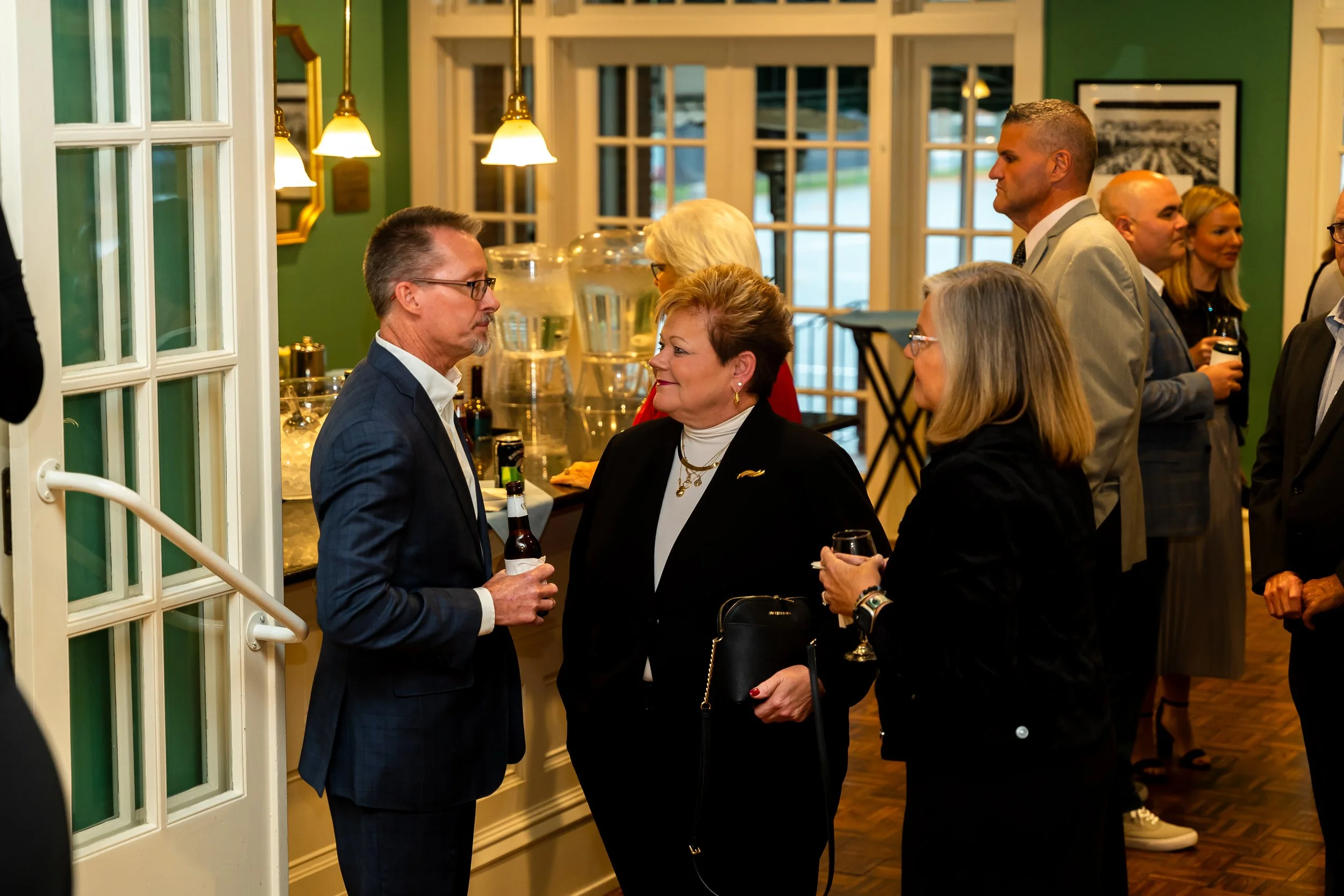 People socializing at an indoor event, engaging in conversation, with drinks in hand, in a well-lit room with large windows and green walls.