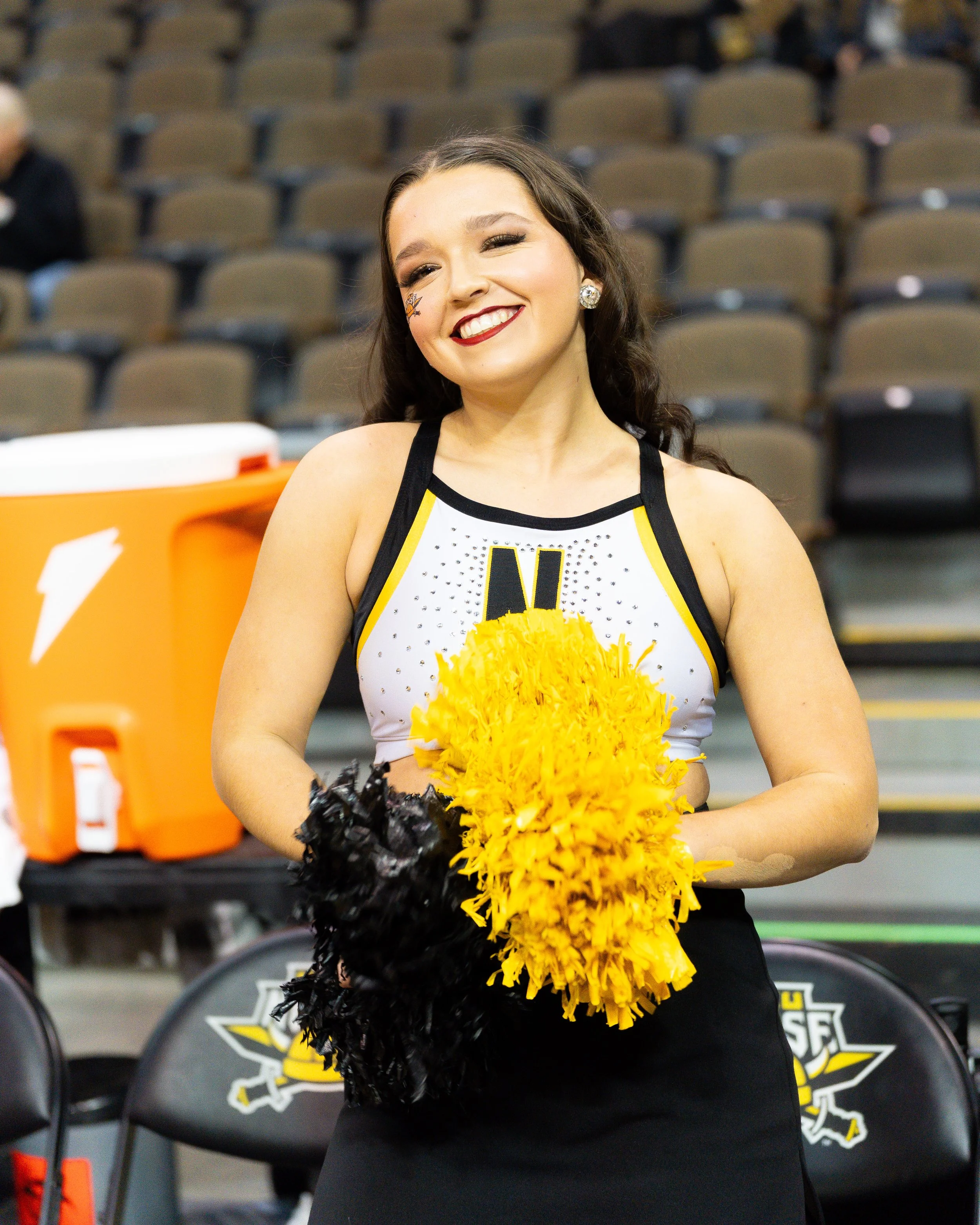A cheerleader in a black and yellow uniform with pom-poms standing on the sideline of a sports arena.