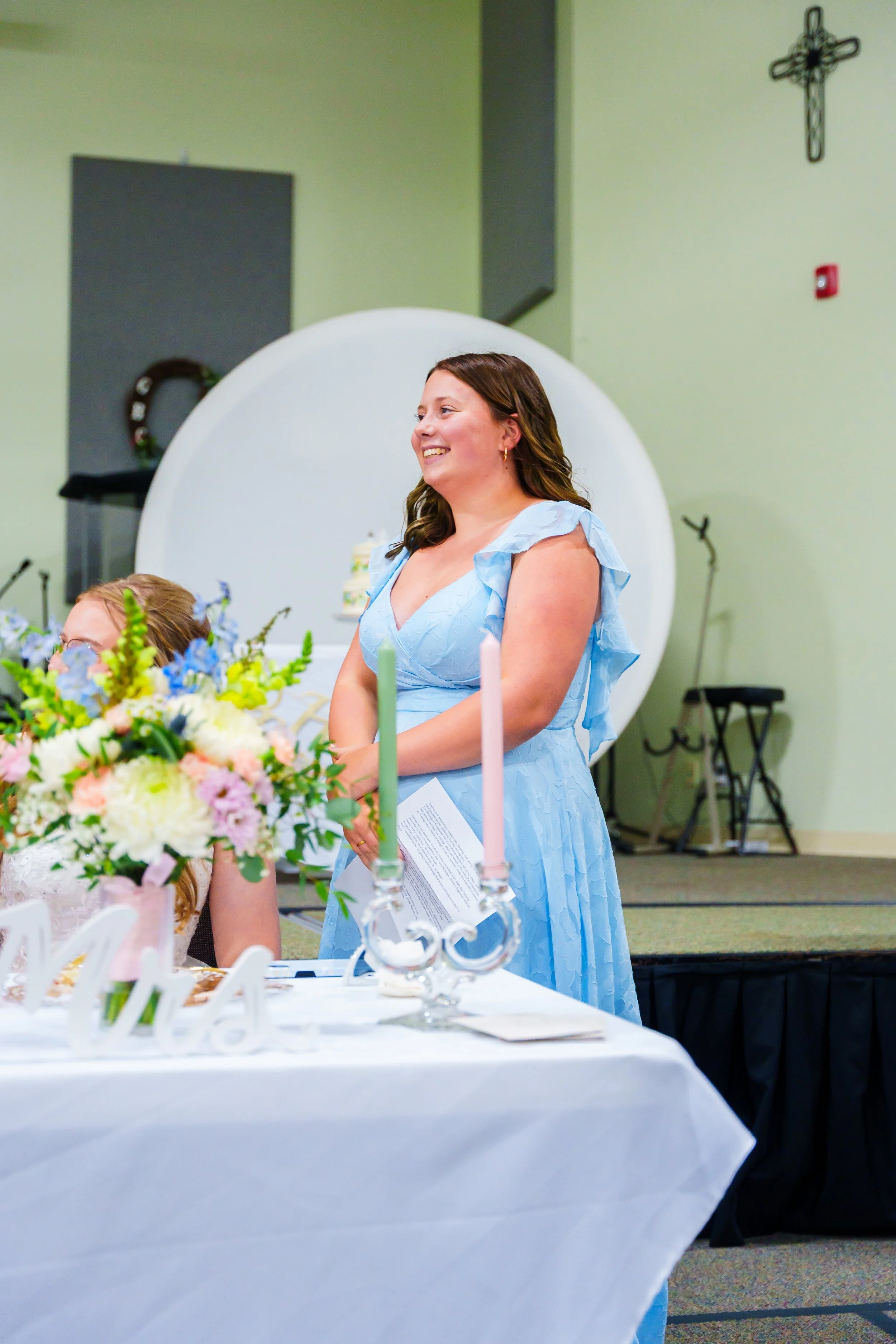 A woman in a light blue dress smiling at a table with floral arrangements and candles during a celebration in a room with church decor.
