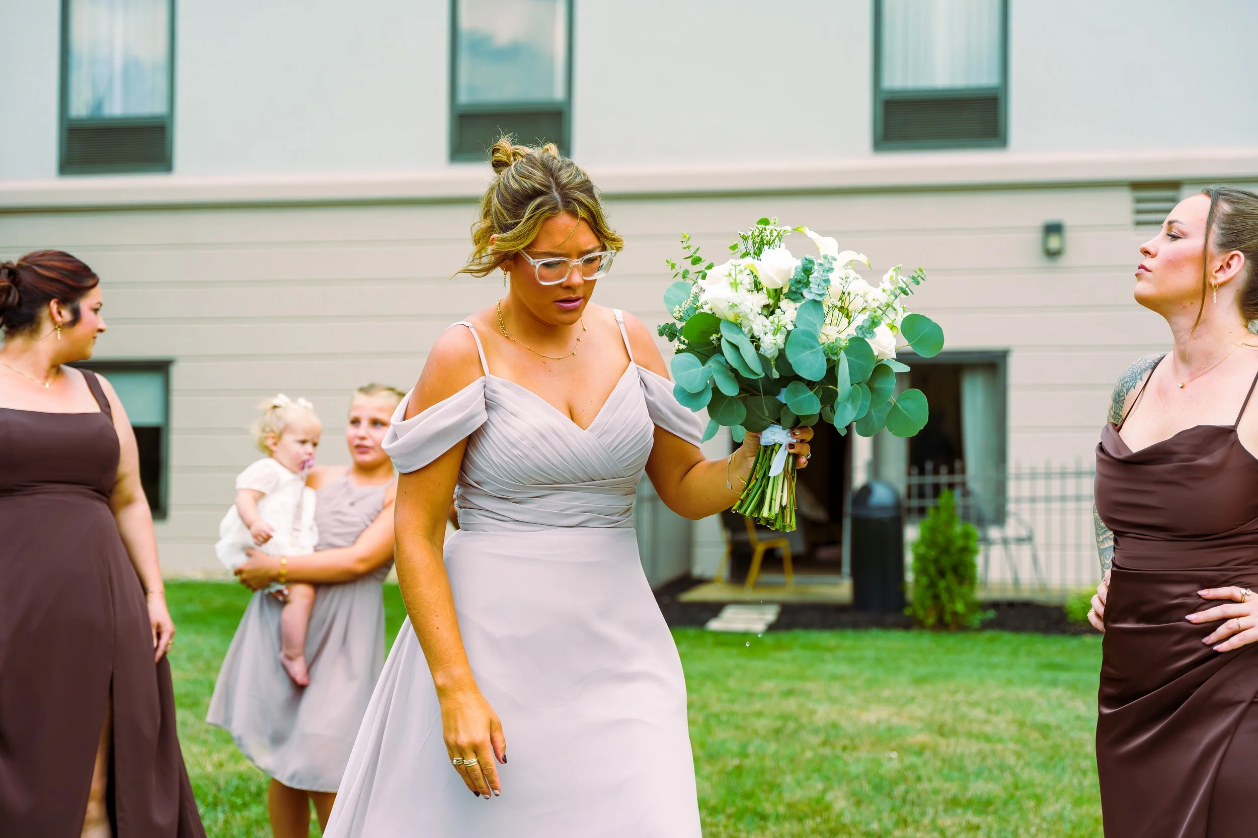 A woman in a light gray dress holding a bouquet of white and green flowers at an outdoor wedding, with other women and children in the background.