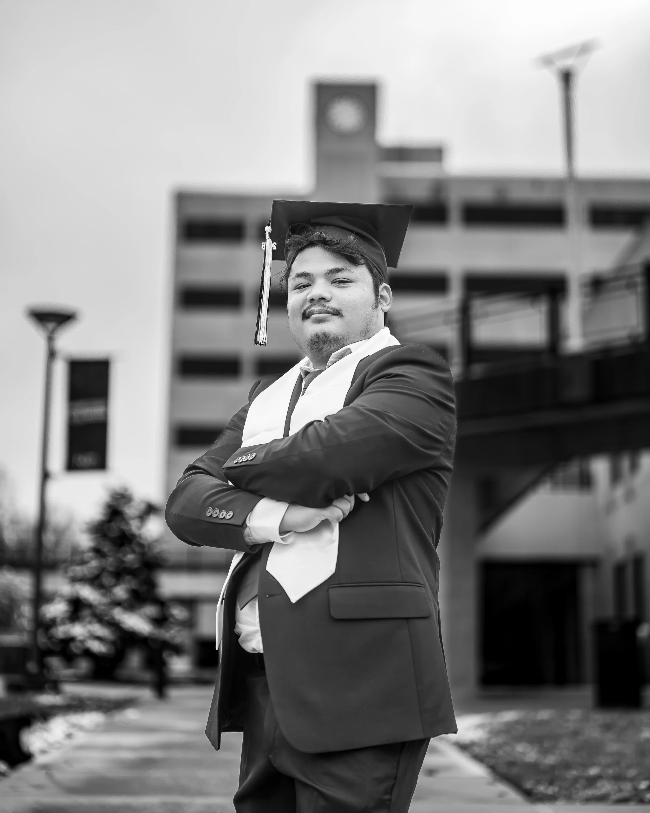 Young man in graduation cap and gown standing outdoors with arms crossed, smiling, with a campus building and clock tower in the background.