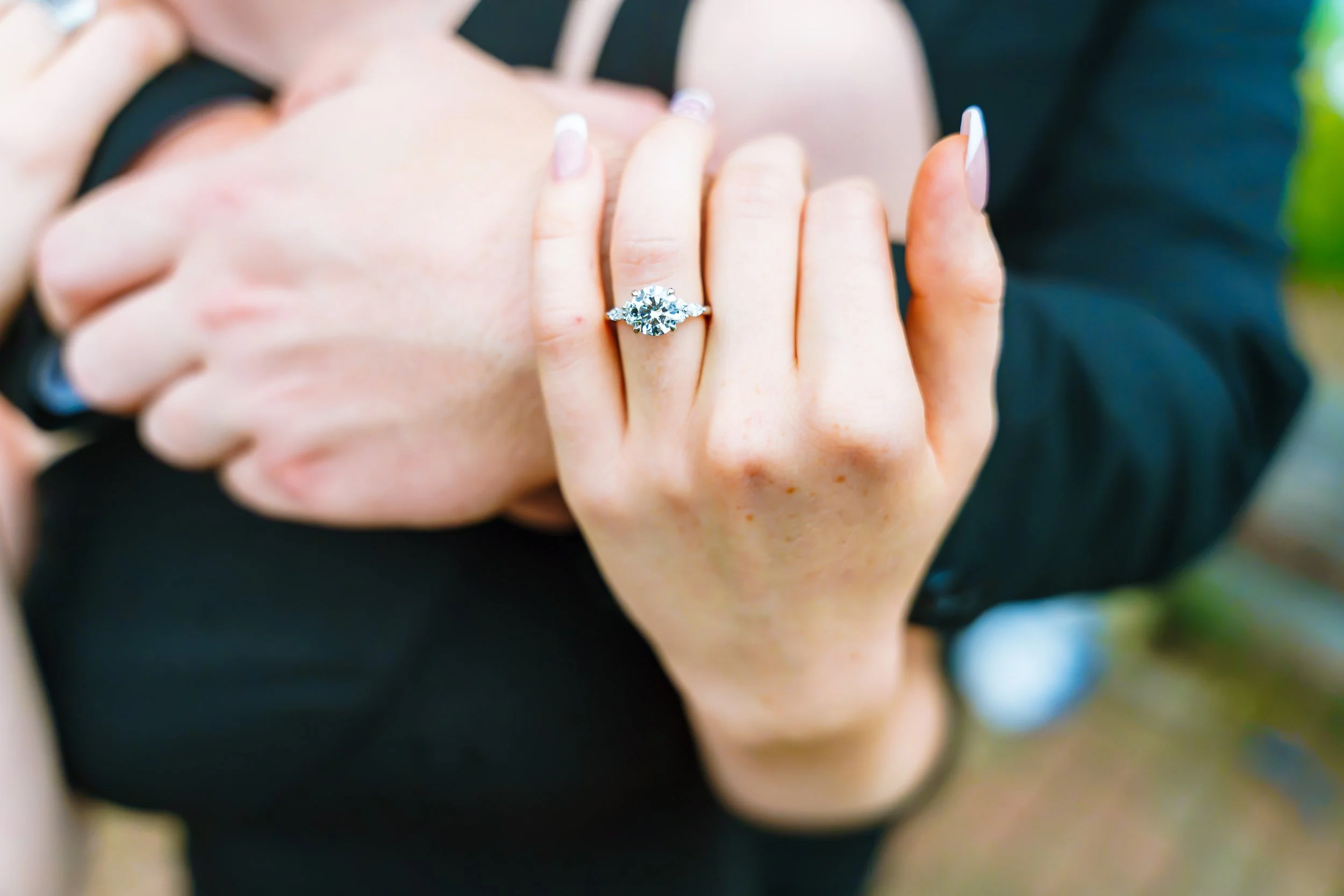 Close-up of a person's hand showing an engagement ring with a large diamond on their finger, with the person wearing a black outfit. The background is blurred with hints of outdoor greenery.