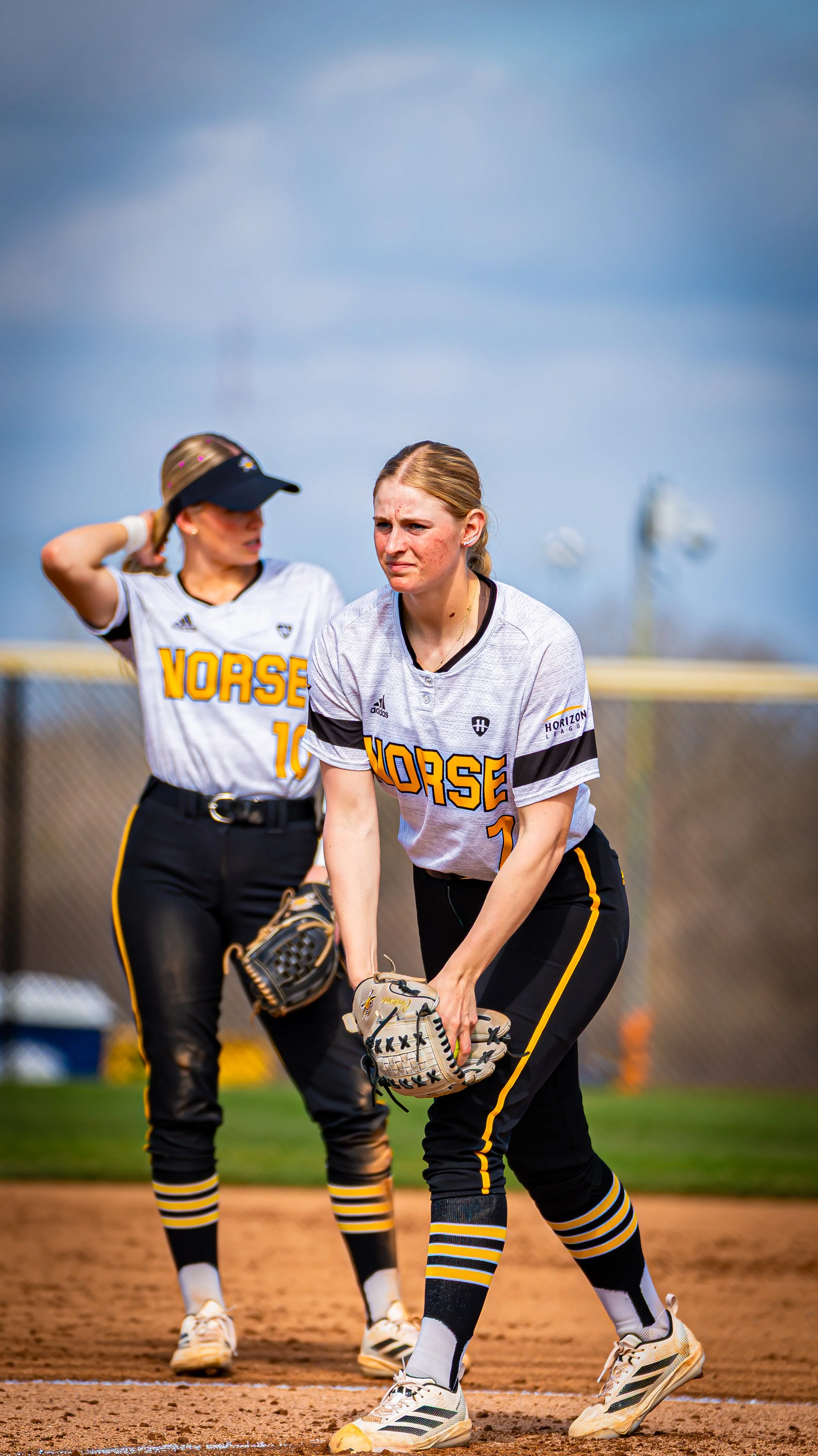 Two female softball players in black and yellow uniforms on the field, one wearing a hat, appear to be preparing for the game.