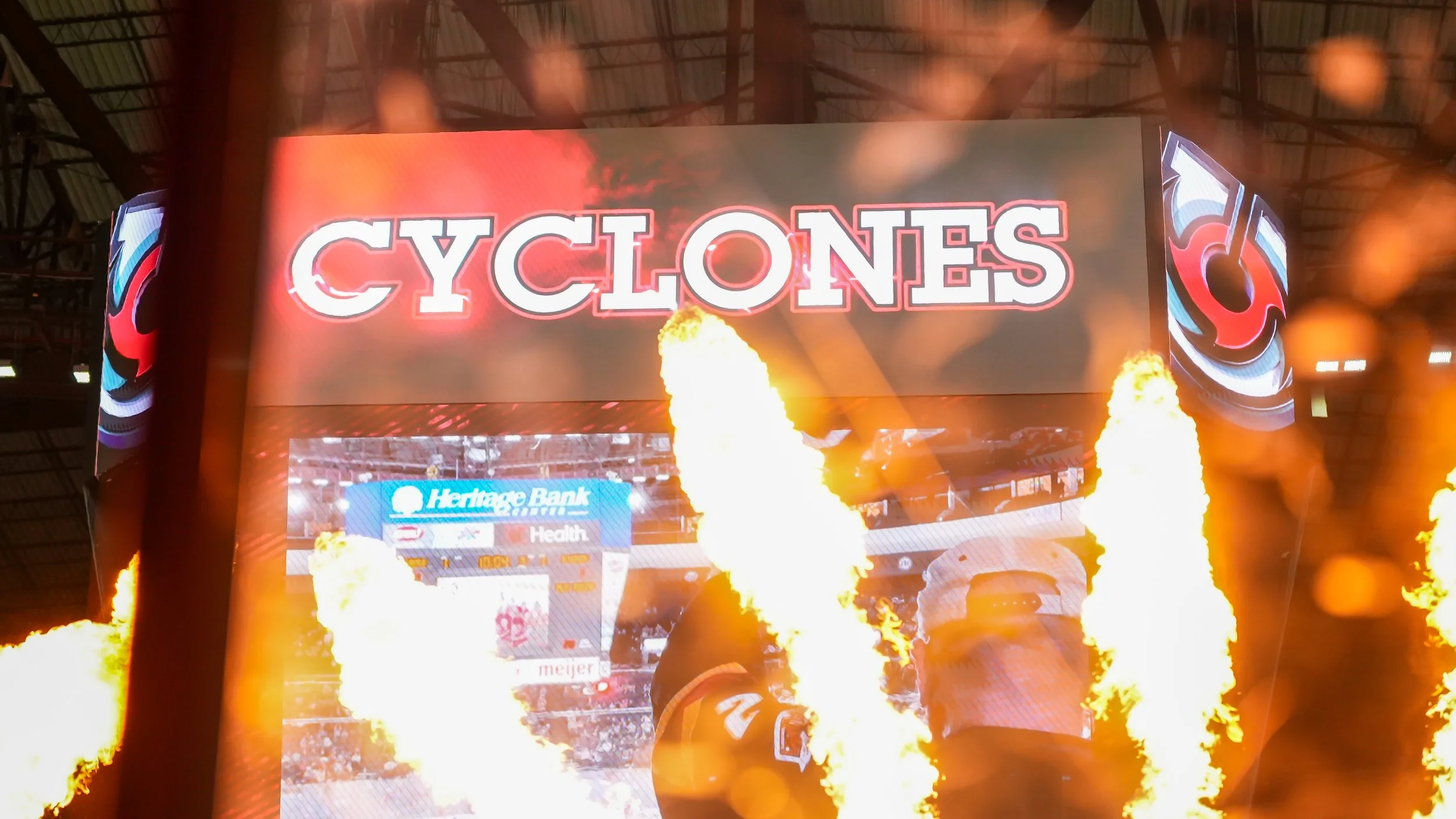 A large digital scoreboard at a sports stadium displays the word 'CYCLONES' with flames and fire effects below it, and parts of the stadium and advertisements are visible in the background.