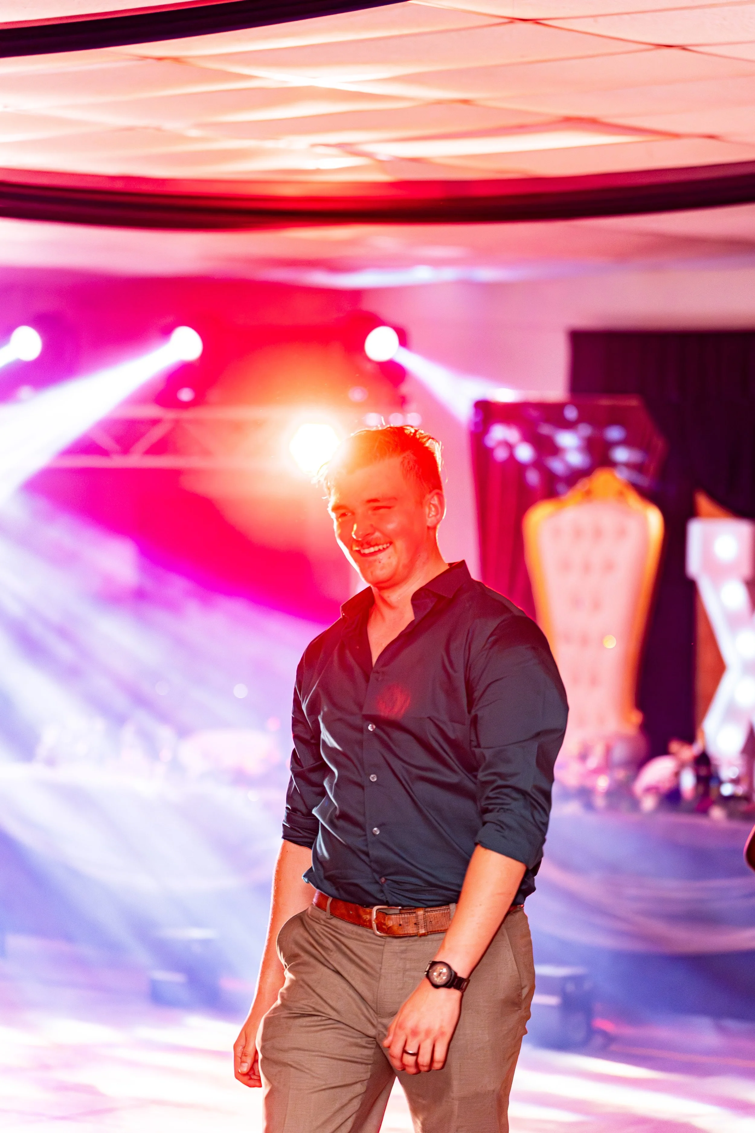 A man in a black shirt and khaki pants standing in front of colorful stage lights and a blurred background at an event or party.