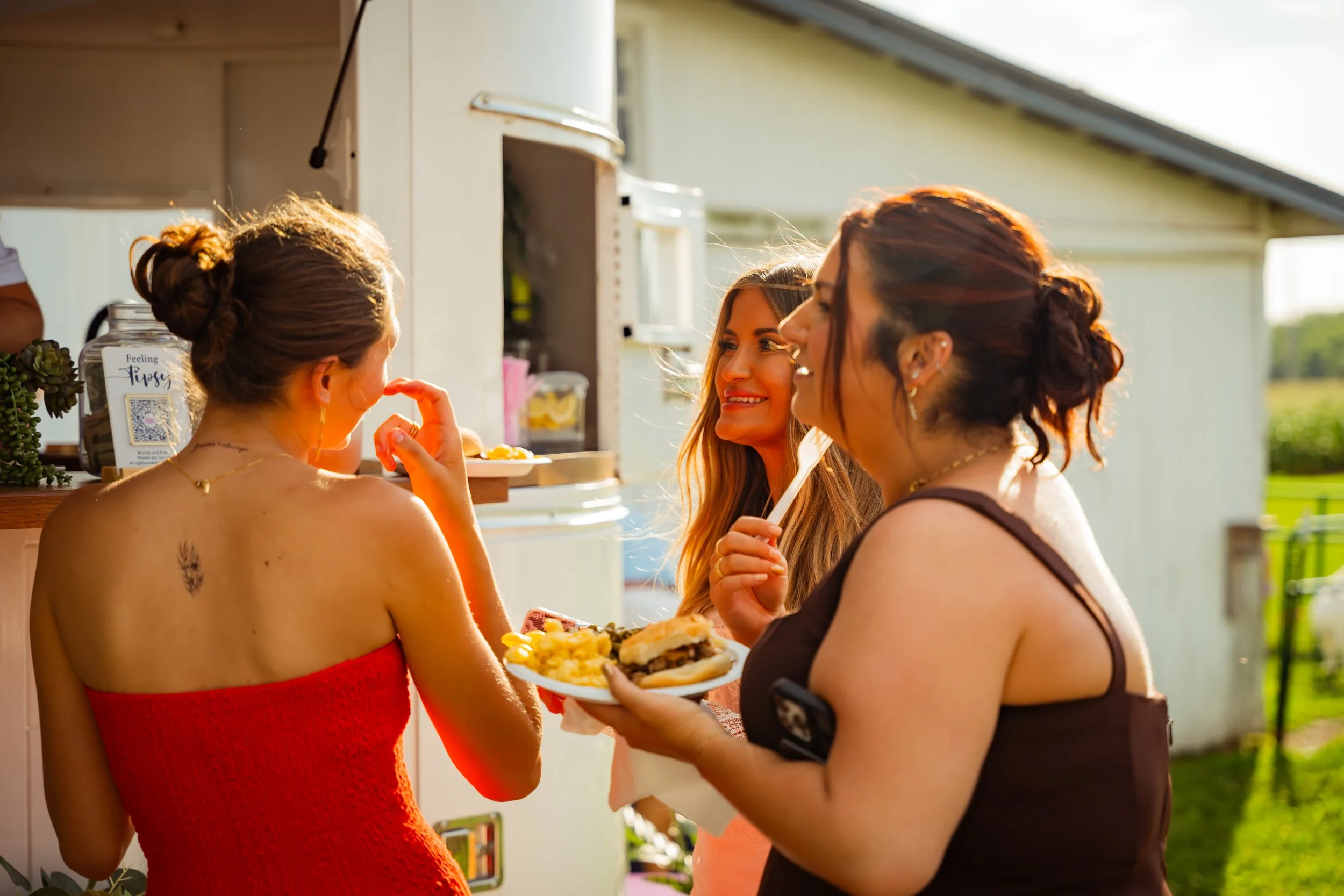 Three women at an outdoor gathering, one holding a plate of food, smiling and engaging in conversation under sunlight.