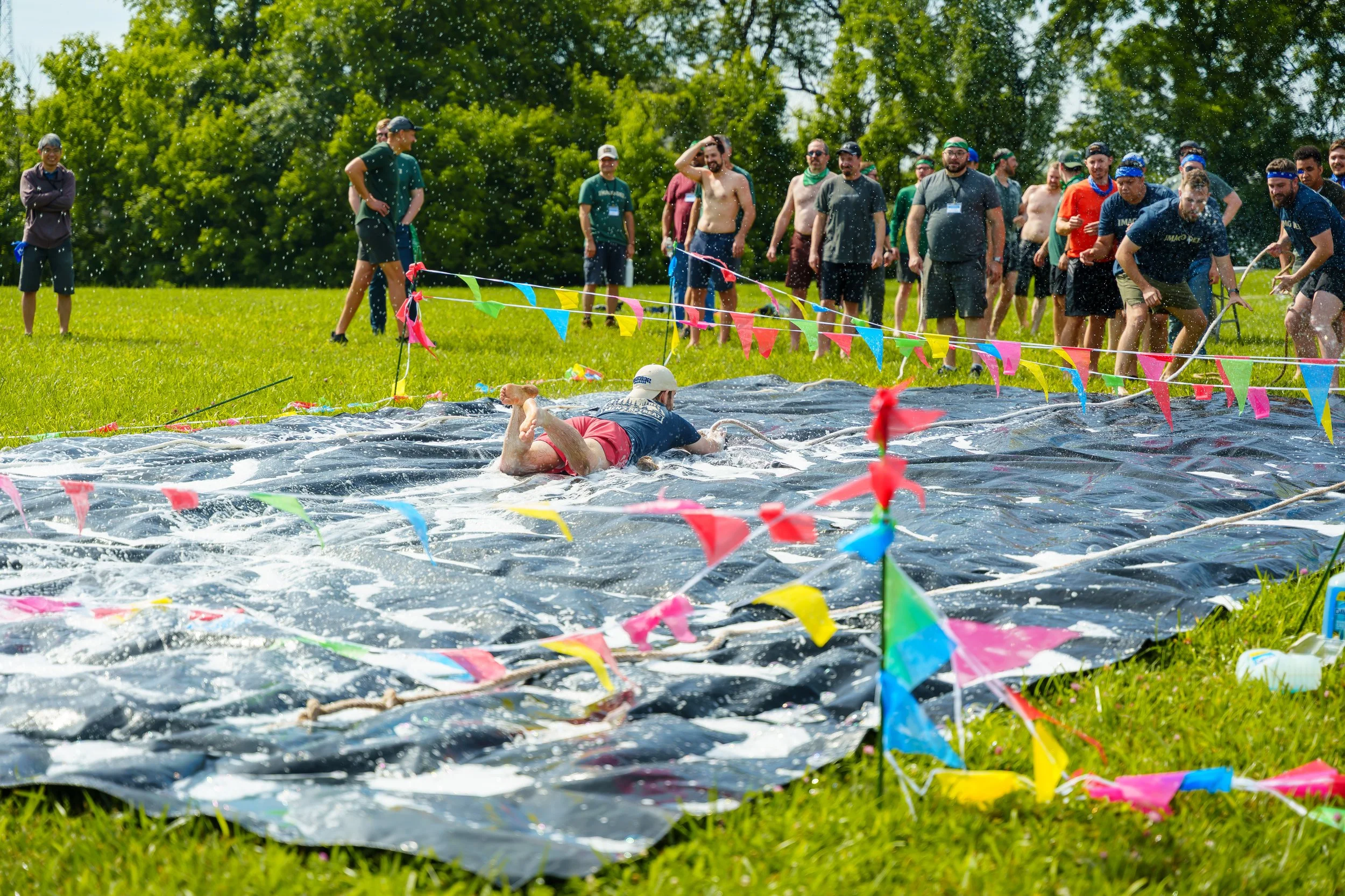 A group of people participating in an outdoor water game on a grassy field, with a person crawling in a water-filled tarp and colorful flags around.