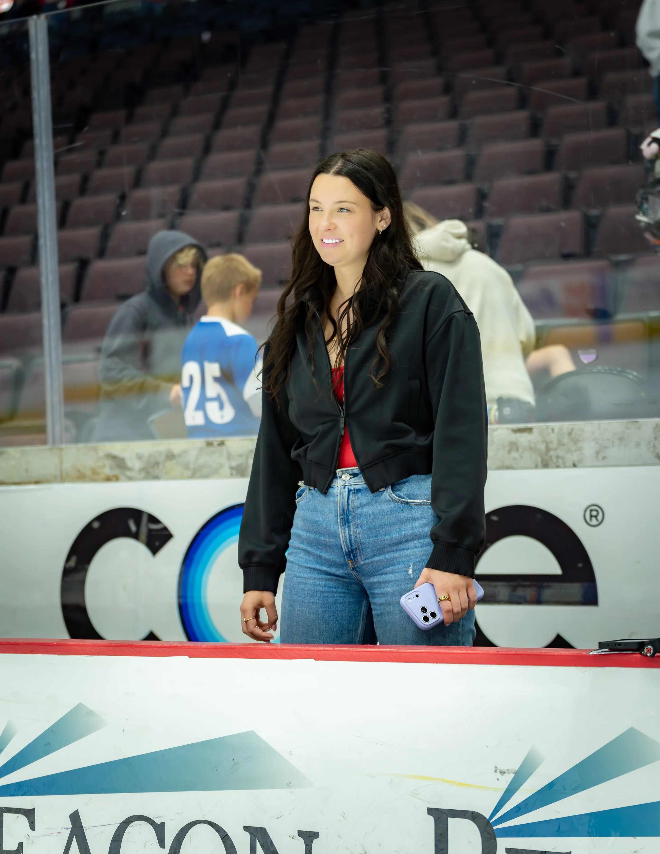 A young woman with long dark hair wearing a black jacket and blue jeans standing at an indoor ice skating rink, holding a smartphone in her right hand, smiling slightly, with other people skating and sitting in the background.