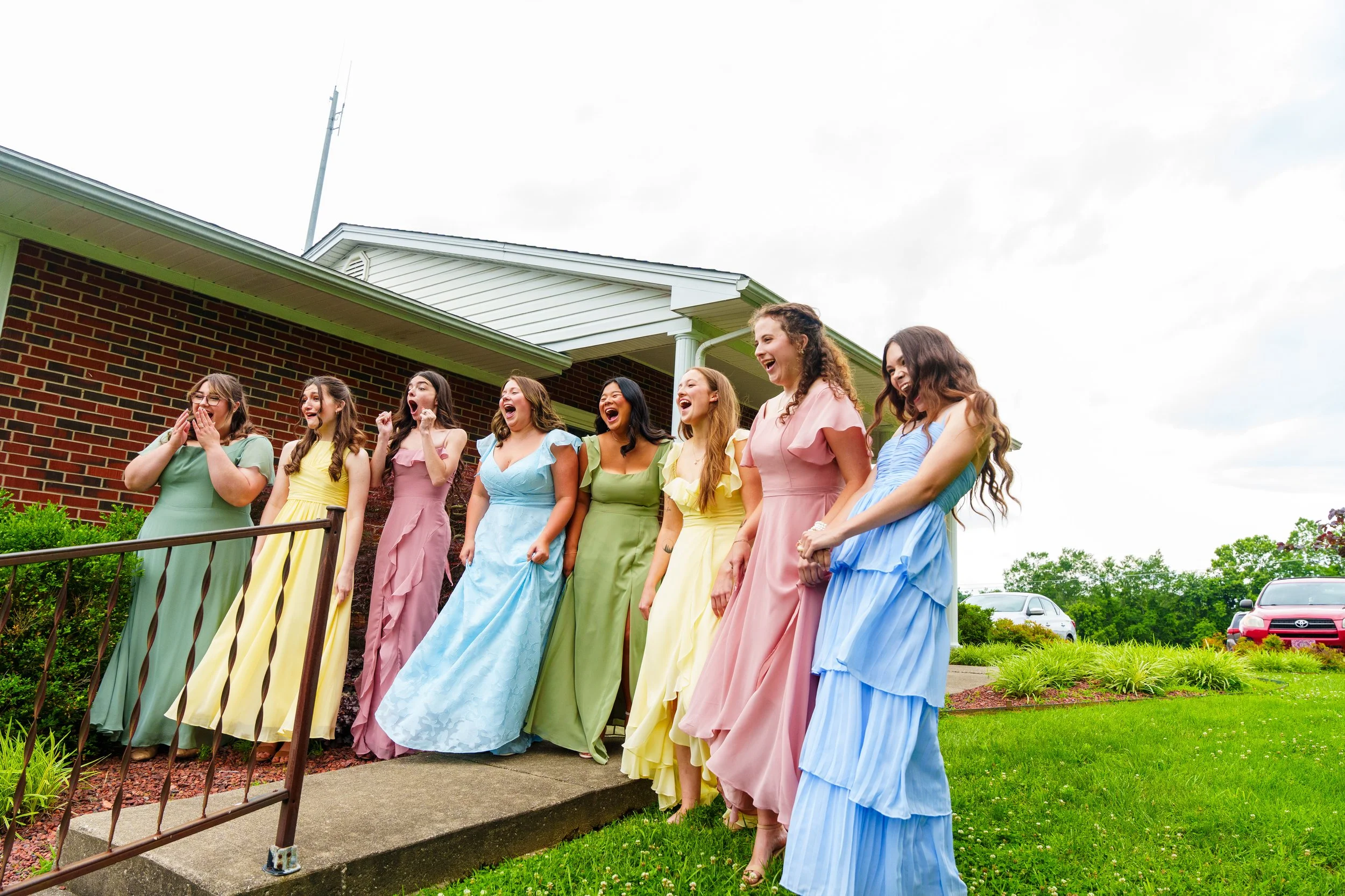 Group of women in pastel-colored dresses standing outside on a sidewalk near a brick building with a green lawn and parked cars, celebrating with joyful expressions.