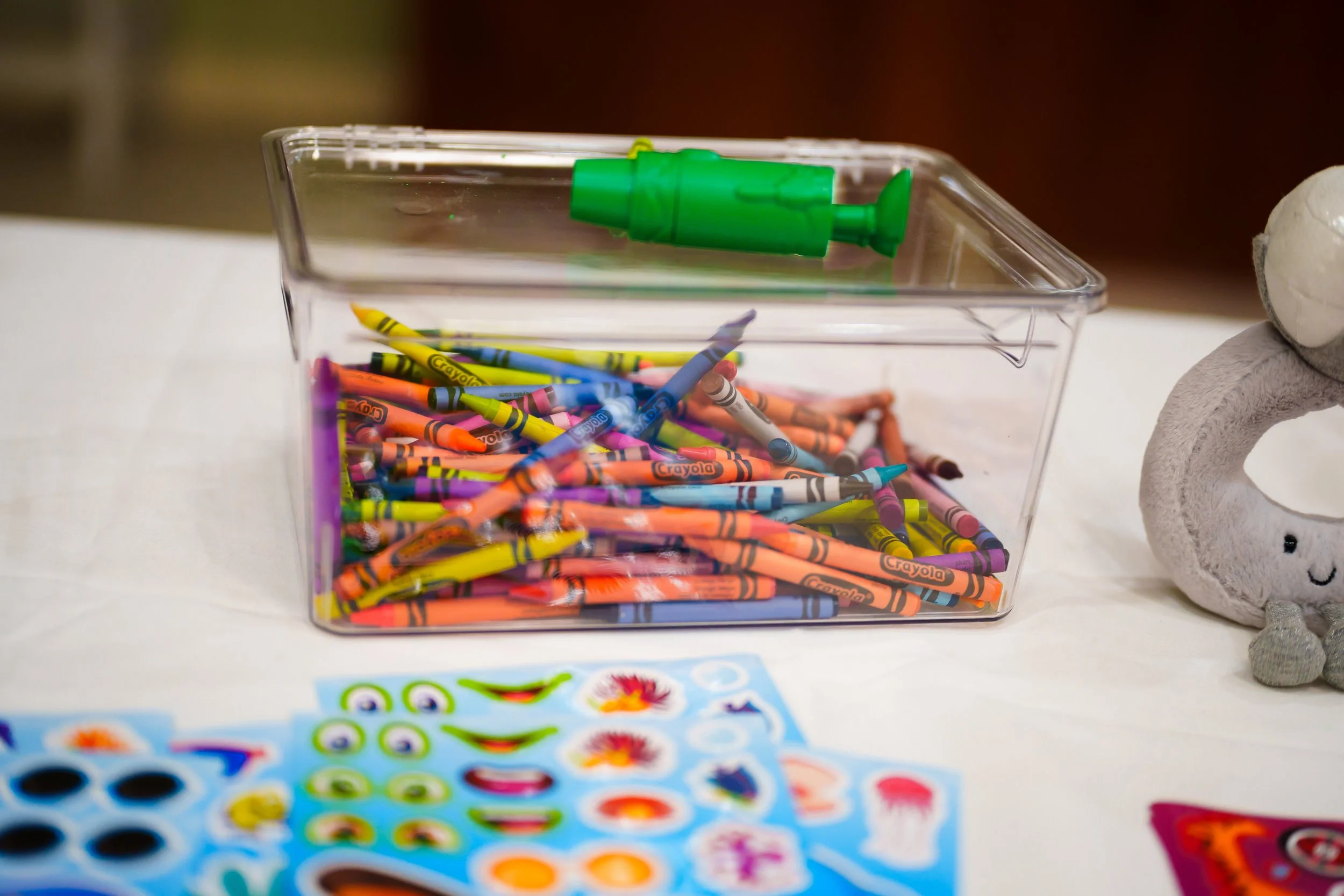 A clear plastic container filled with colorful crayon sticks, with a green glue bottle on top and a gray plush toy with robot-like features on the right side.