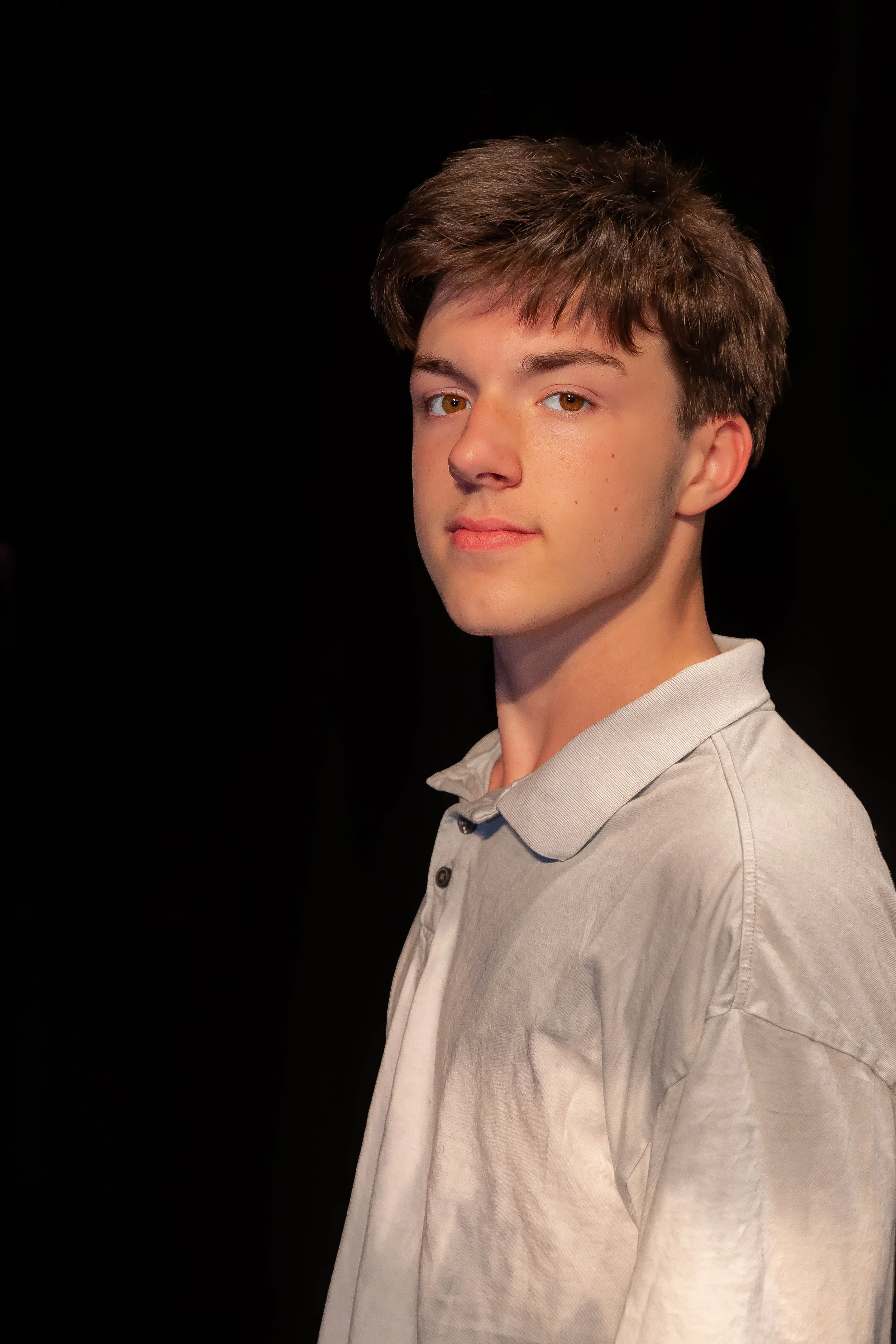 A young man with brown hair and light skin, wearing a light grey button-up shirt, is posing against a black background. He is looking slightly to the left with a neutral expression.