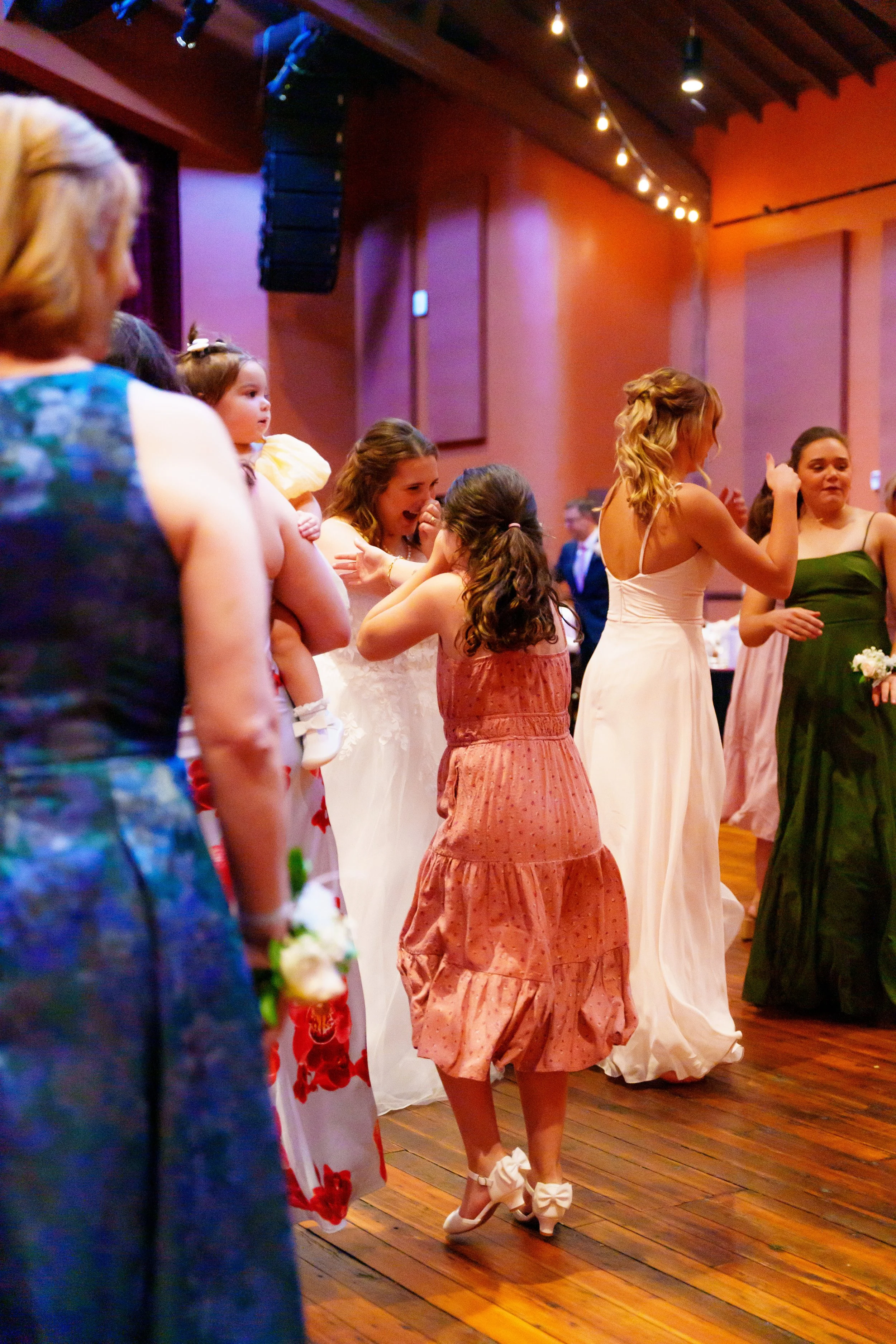 Group of women and girls dressed in fancy dresses, standing on a wooden dance floor at a party or celebration.