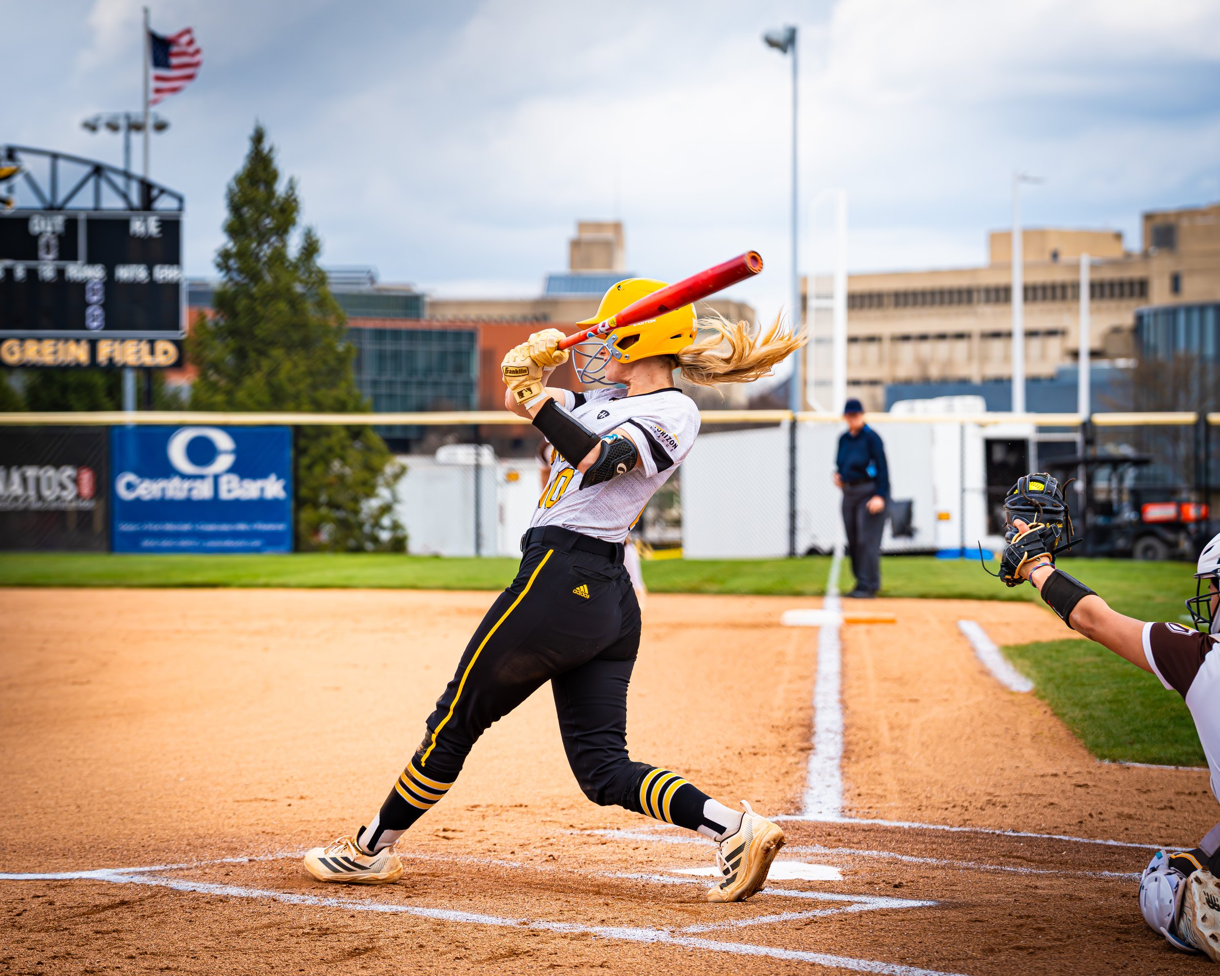 A female softball player in uniform batting during a game, with a cityscape and scoreboard in the background.