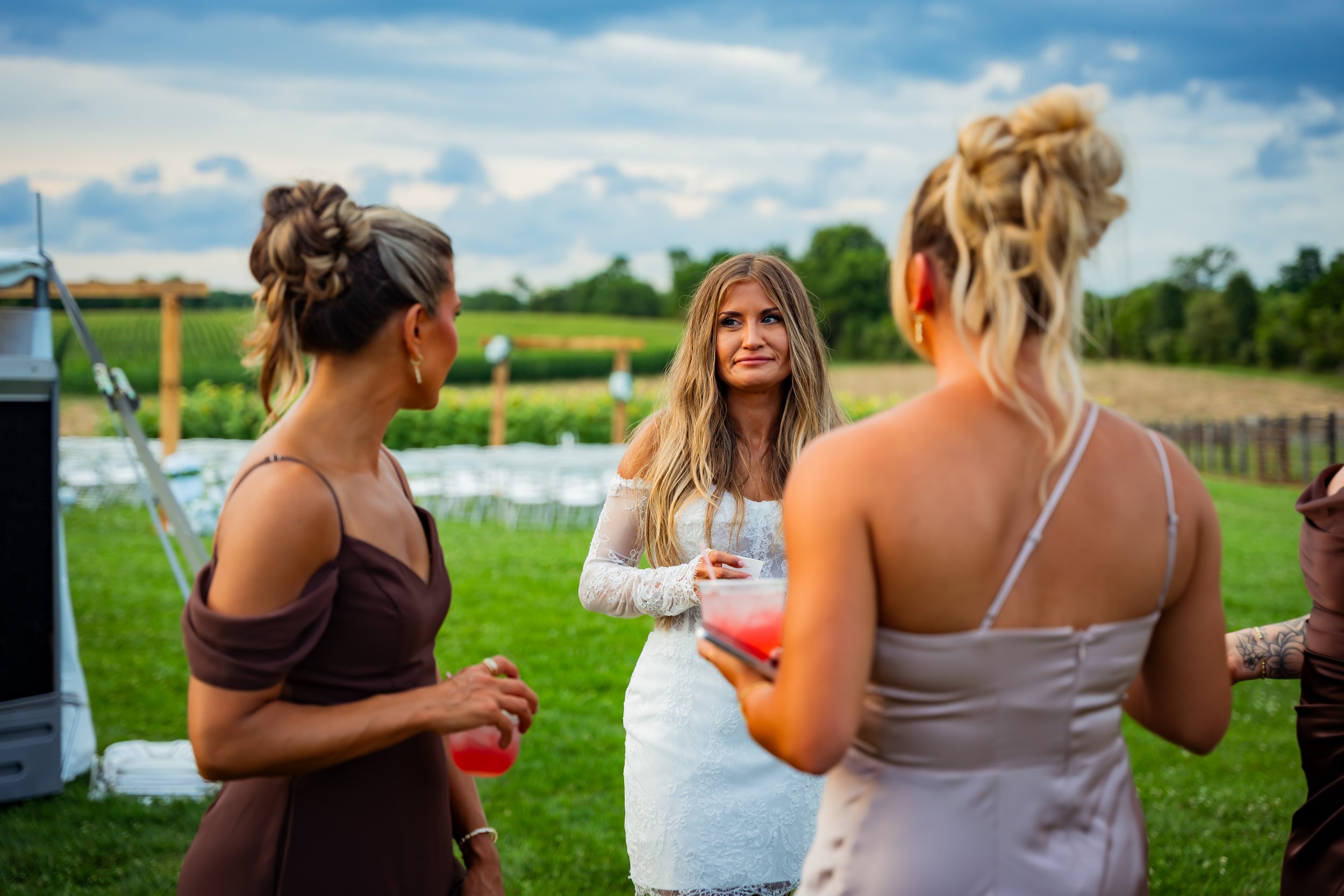 Three women at an outdoor gathering, one in a white dress with lace details and two in brown tops, engaging in conversation with a woman in a white dress, while holding drinks, in a green field under a cloudy sky.