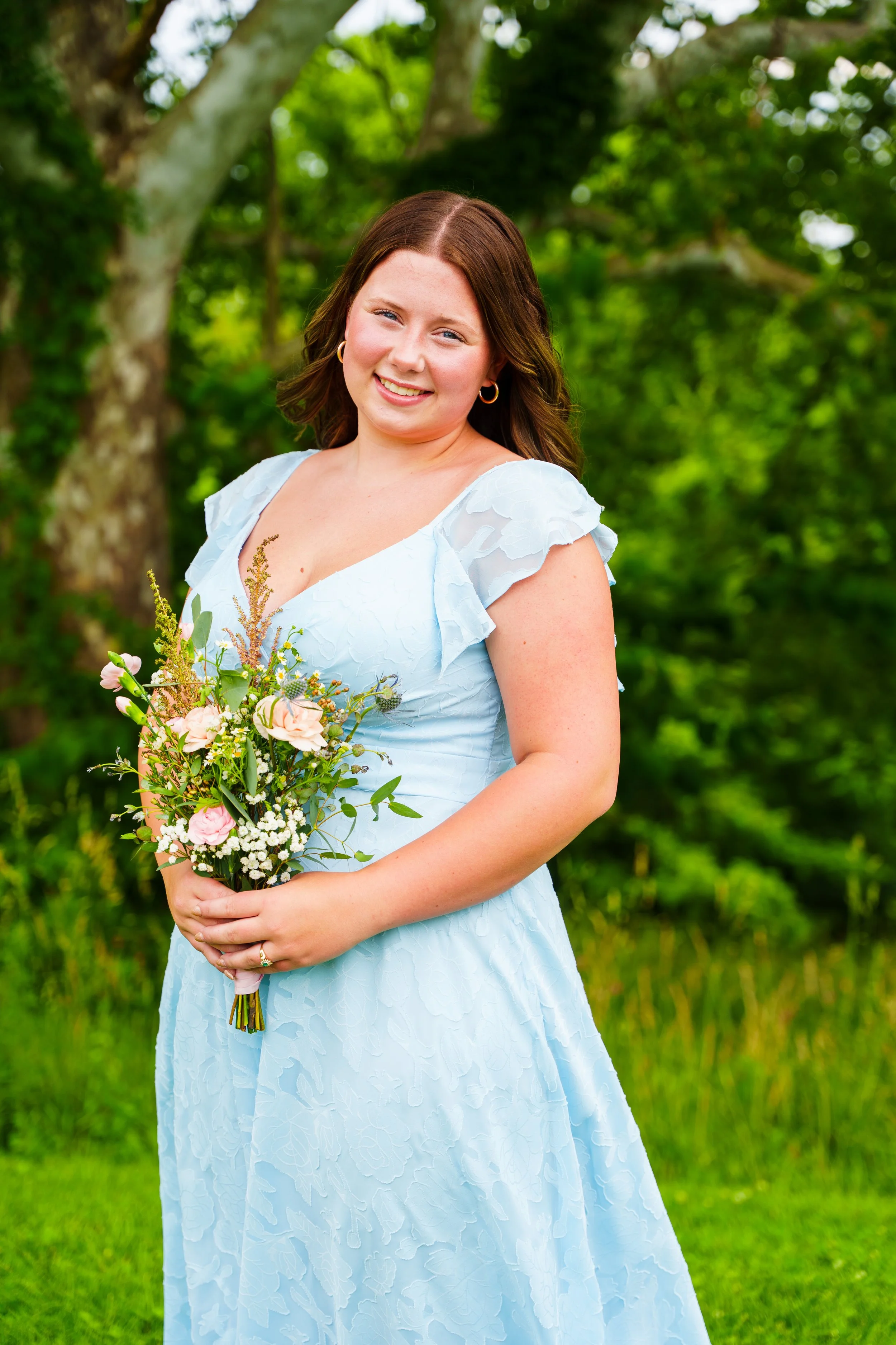 A young woman with brown hair and blue eyes standing outdoors in a green setting, wearing a light blue dress with ruffled sleeves, holding a bouquet of pink and white flowers with greenery.