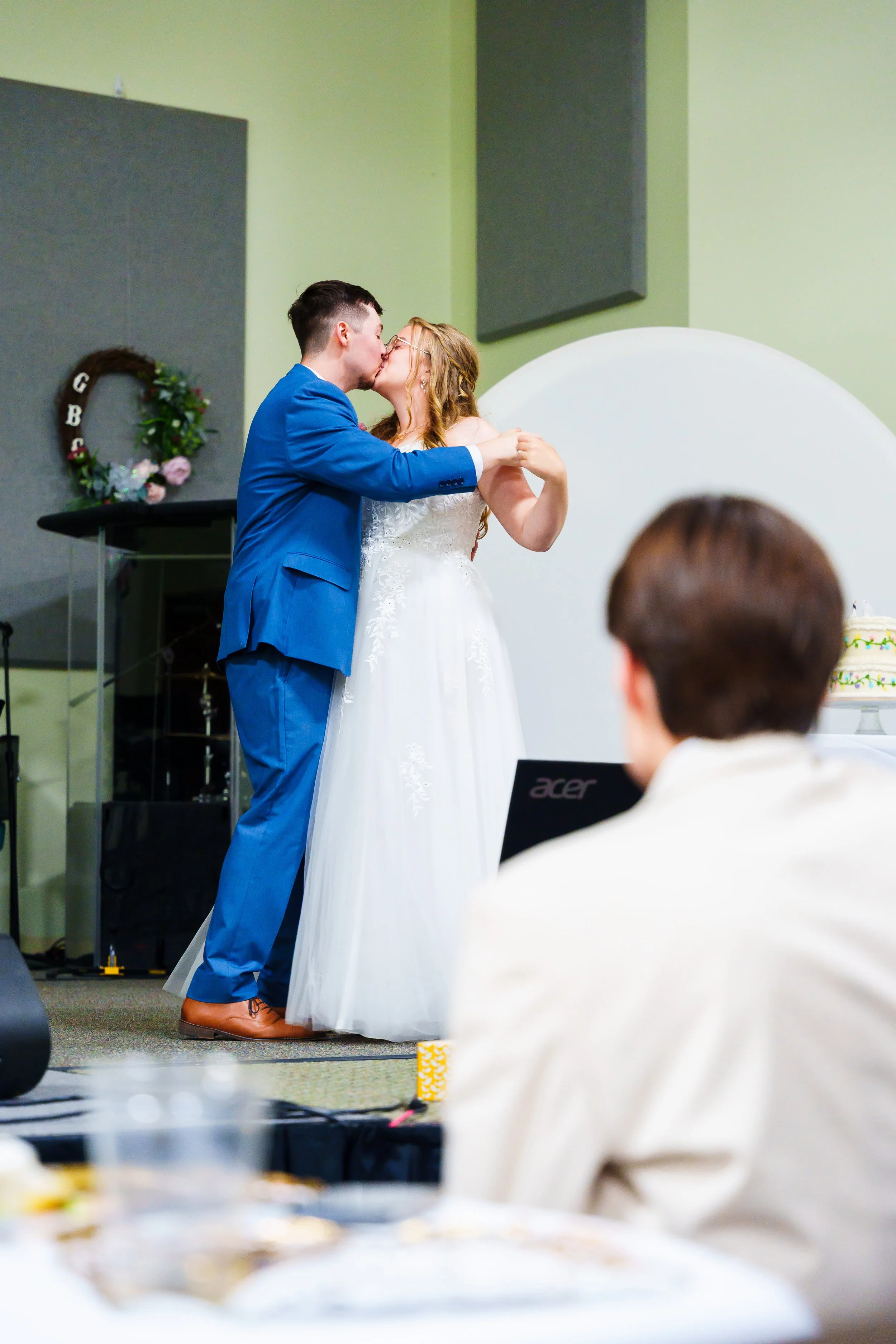 A couple dressed in wedding attire sharing a kiss at a wedding ceremony. The groom wears a bright blue suit, and the bride wears a white wedding dress. People are seated and watching, with a decorated wedding wreath hanging on the wall in the backgro