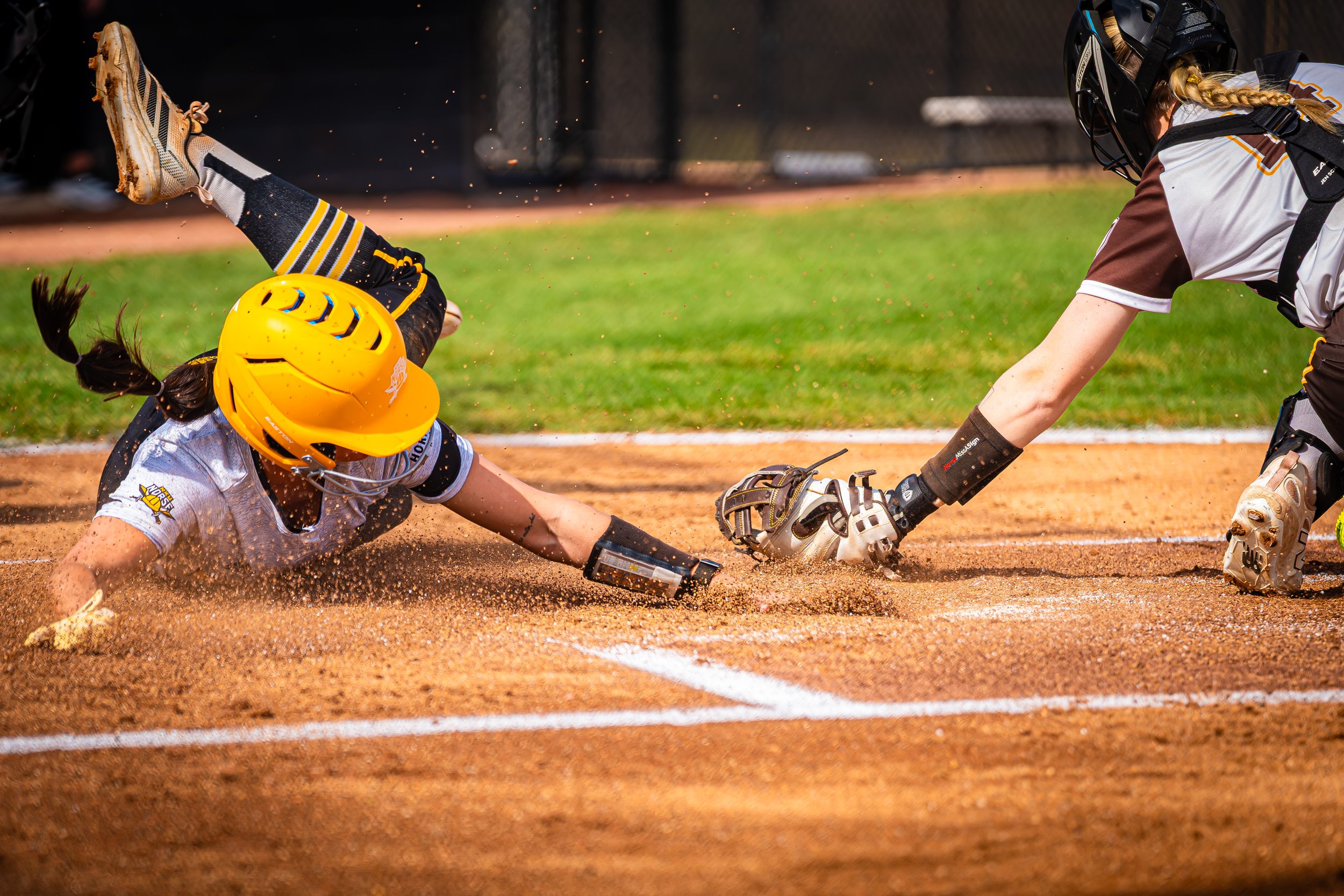 A youth baseball player wearing a yellow helmet and white uniform sliding into home plate while another player in a gray and black uniform tries to tag him out on a dirt baseball field.