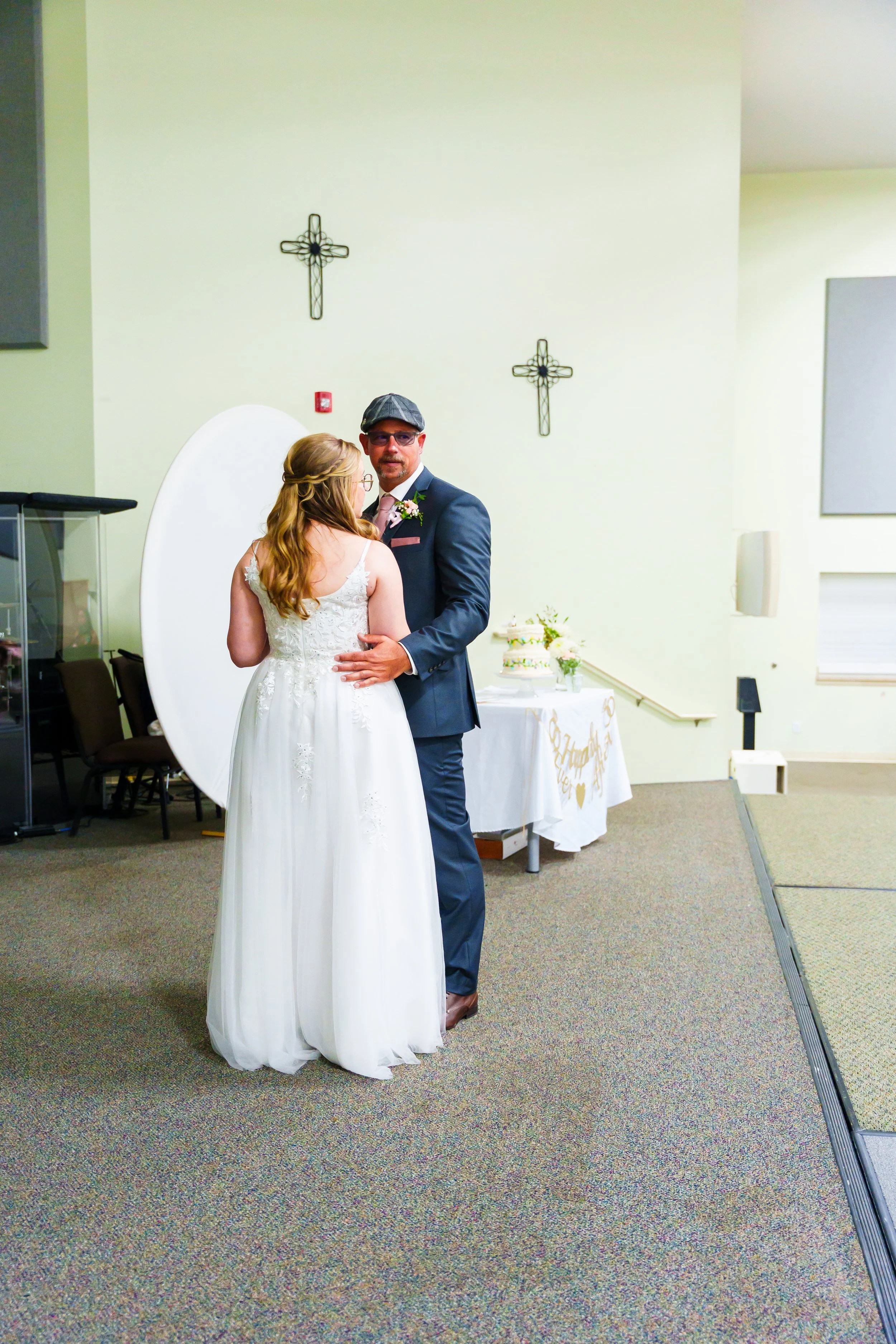 A bride and groom sharing a dance at their wedding reception. The bride is wearing a white wedding gown, and the groom is in a dark suit with a hat. They are standing in a room decorated with crosses on the wall and a table with a cake and flowers in