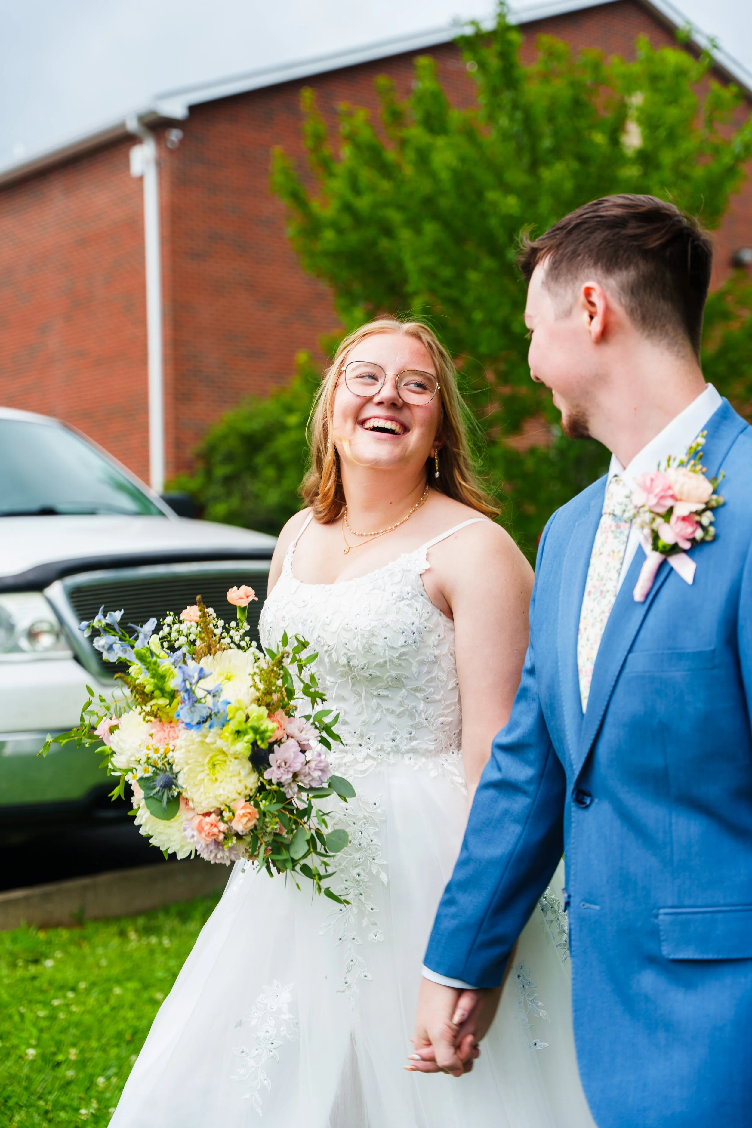 A bride and groom holding hands at a wedding outdoors. The bride is smiling at the groom, wearing glasses and a white wedding dress, holding a large bouquet of flowers. The groom is wearing a blue suit with a floral tie and boutonniere. They are in f