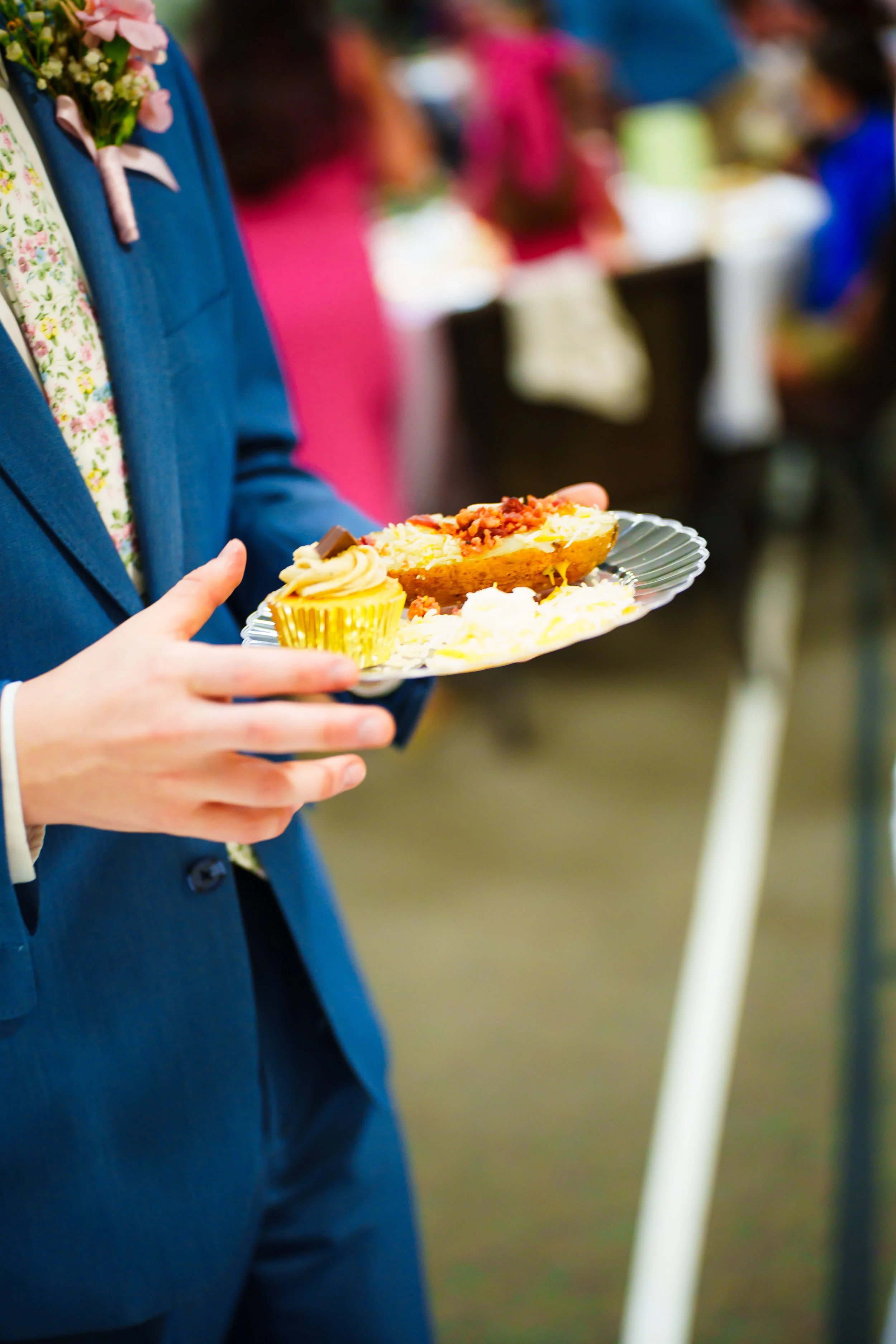 Person in blue suit holding a plate of food at a social event, with people in colorful clothing in the background.