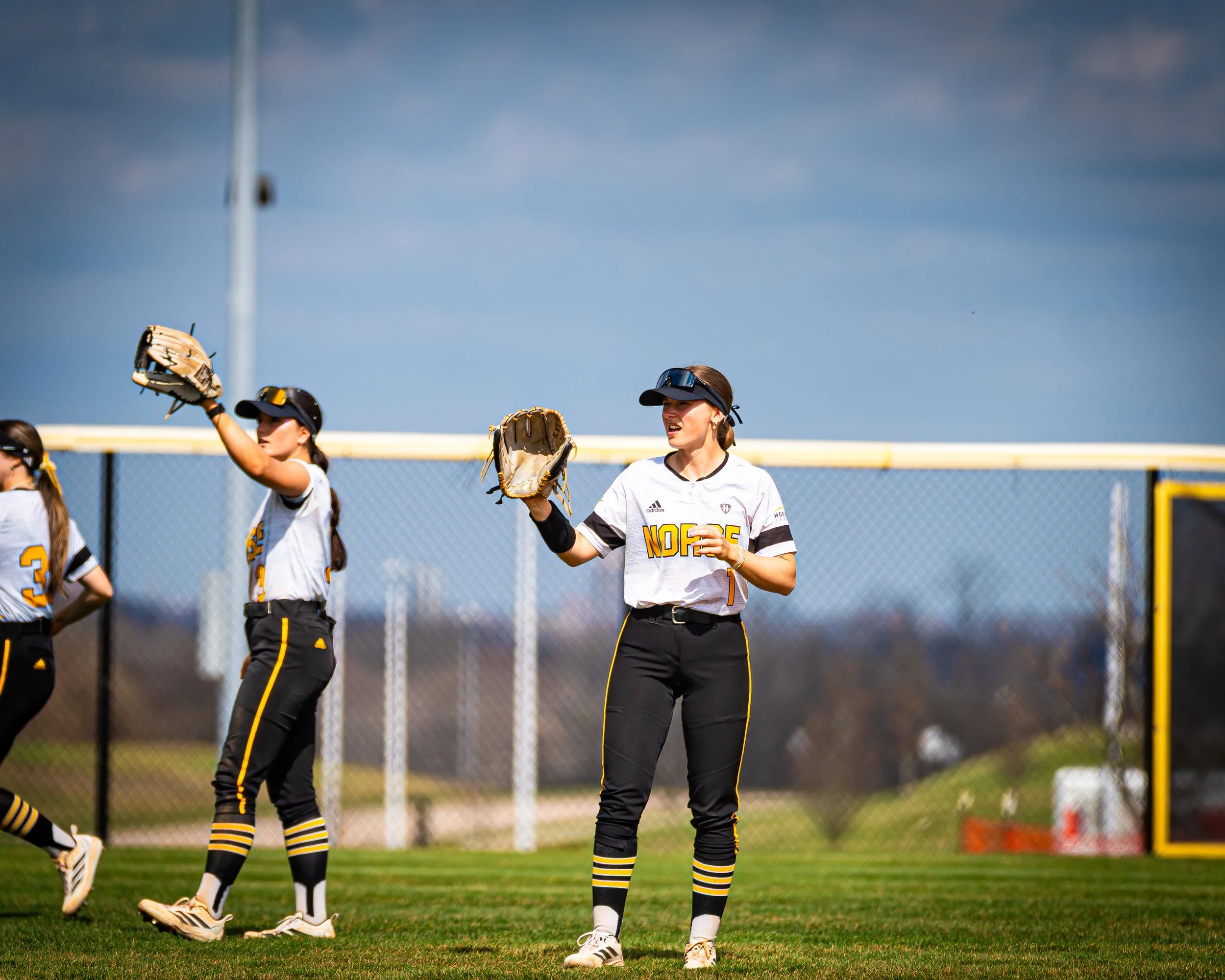 Girls softball players wearing black and yellow uniforms, some wearing visors, standing on a grassy field with a chain-link fence in the background, preparing for the game.