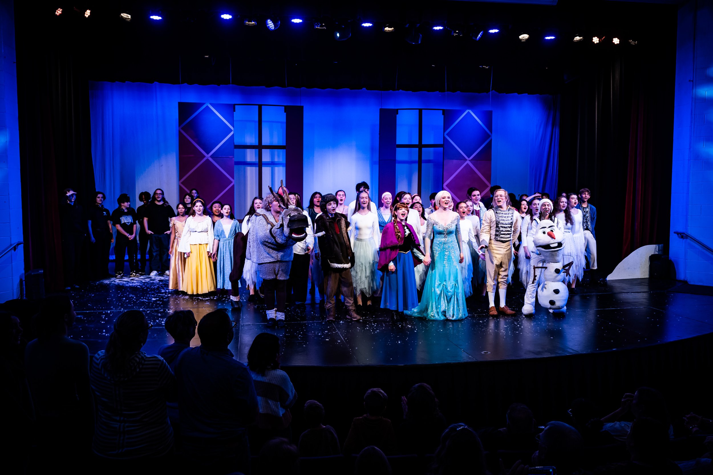Stage performance with children in costumes, including characters from Frozen, with a backdrop of blue curtains and windows, and an audience watching.