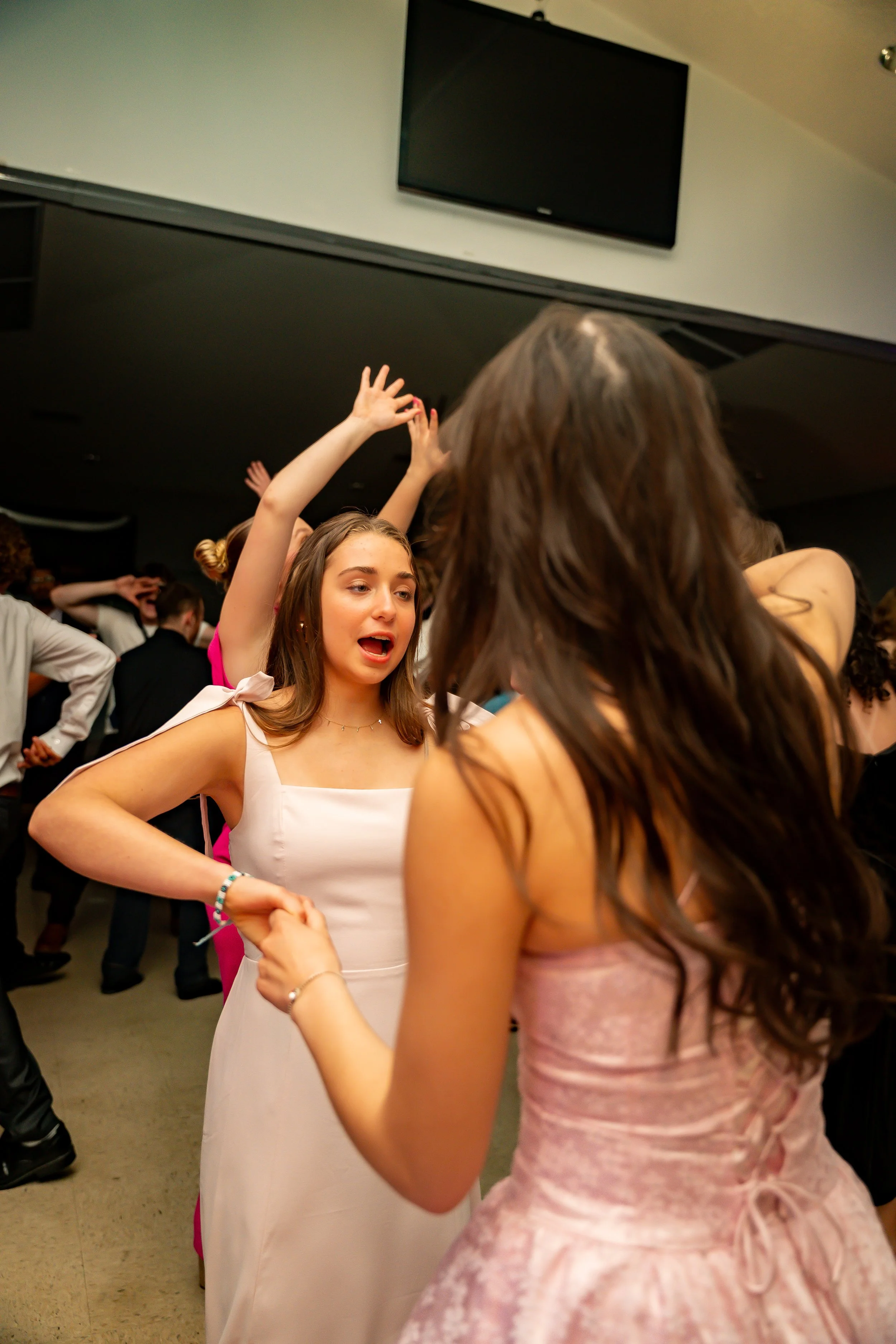 A group of young women dancing and having fun at a party or celebration indoors. One woman in a white dress is smiling and raising her arms, while another woman in a pink dress is seen from behind, holding hands with her.