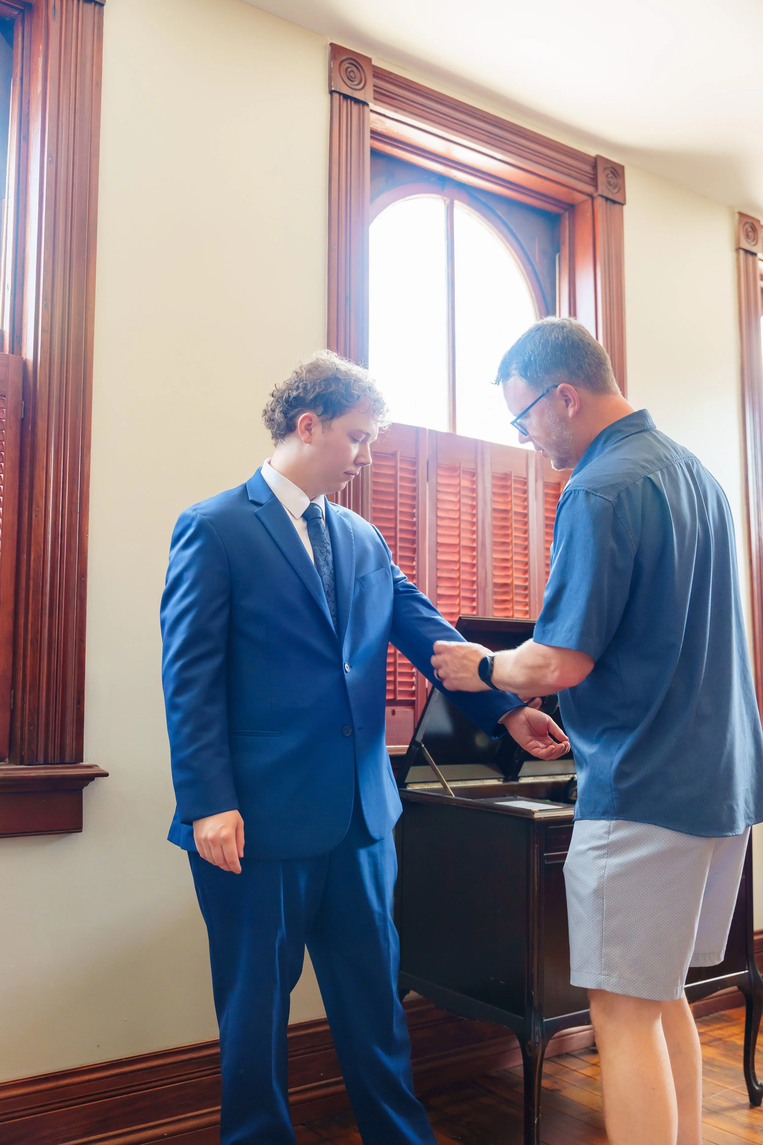 A young man in a blue suit and tie is standing in a room with wooden window frames and wood-paneled walls, receiving something from an older man in a casual blue shirt and shorts near a piano.