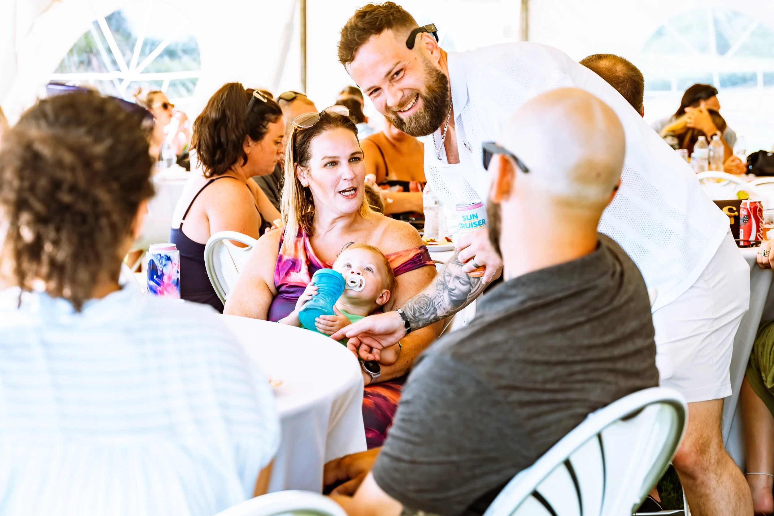 People gathered at a social event, sitting at tables under a tent, engaging in conversation. One man is smiling and leaning over to talk to women, including a woman holding a baby.