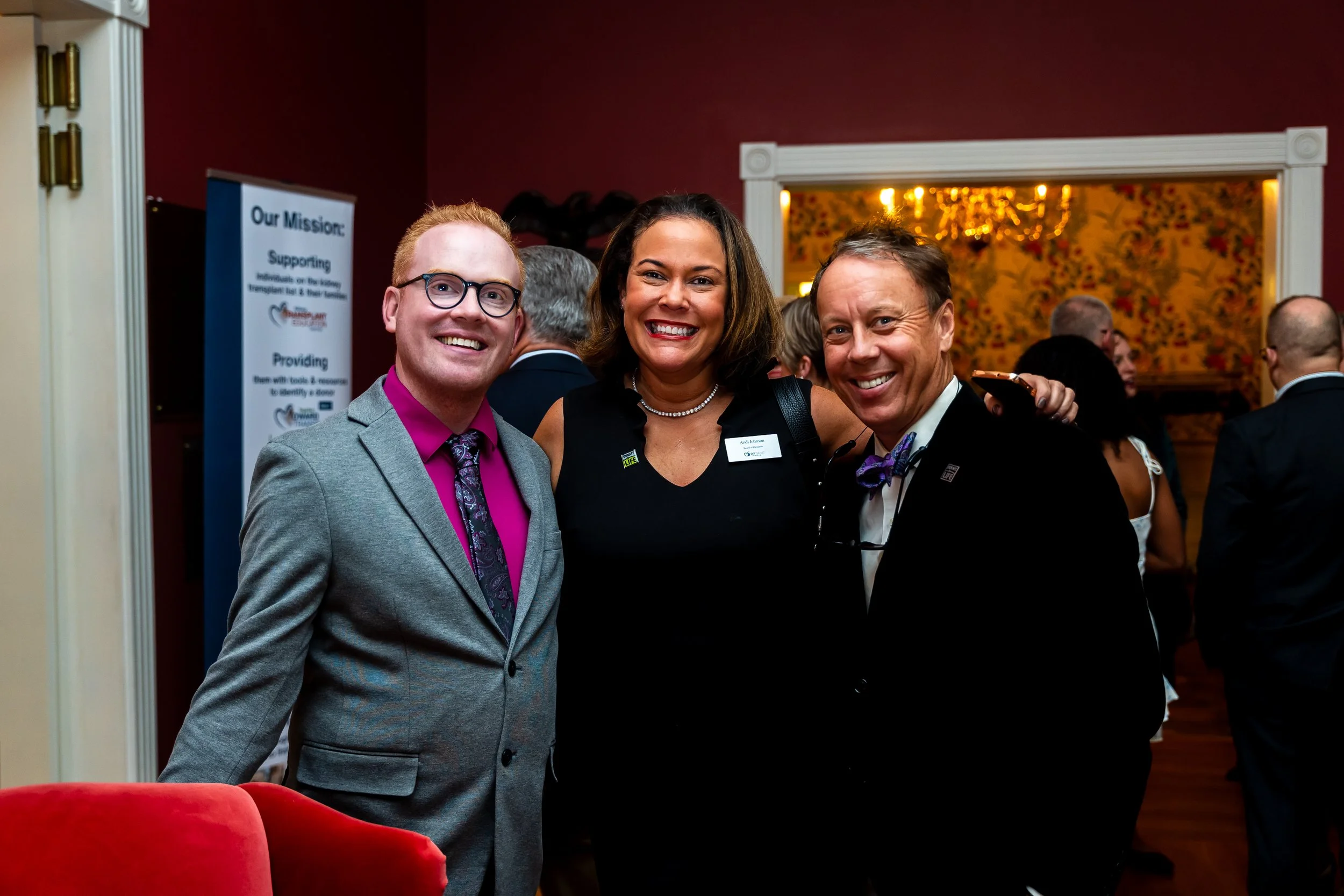 Three people smiling and posing for a photo at a formal event, with a red wall and a floral-patterned background.