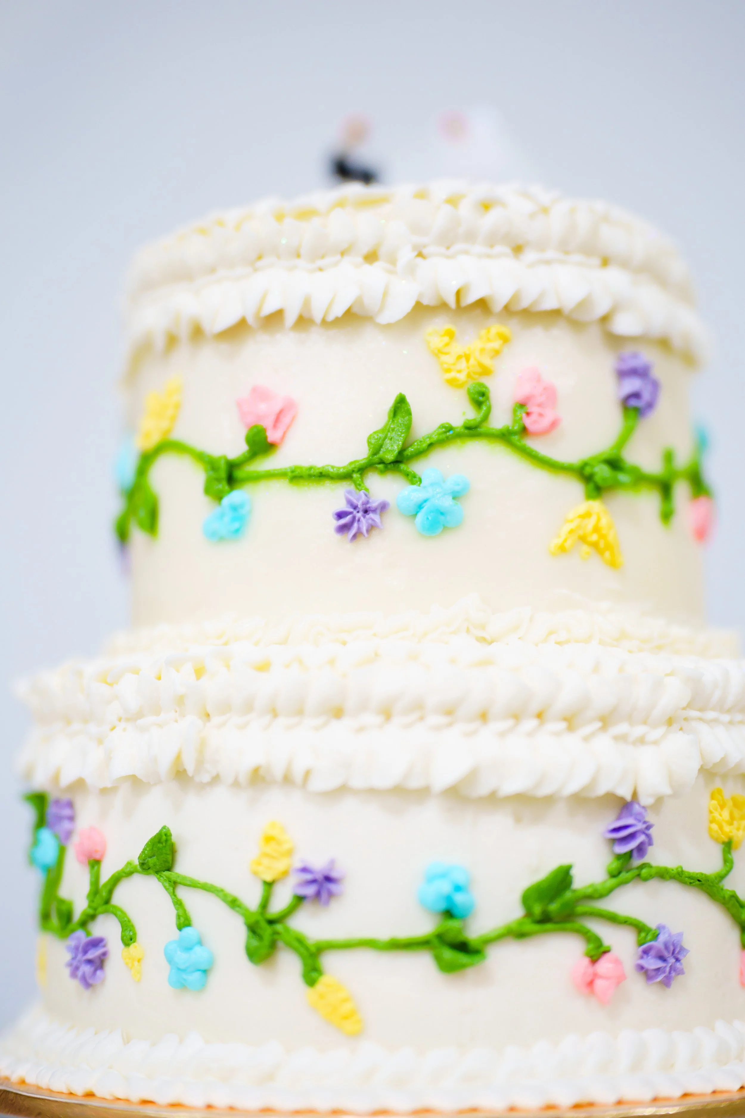 Close-up of a white layered cake decorated with colorful piped icing flowers and green vines.