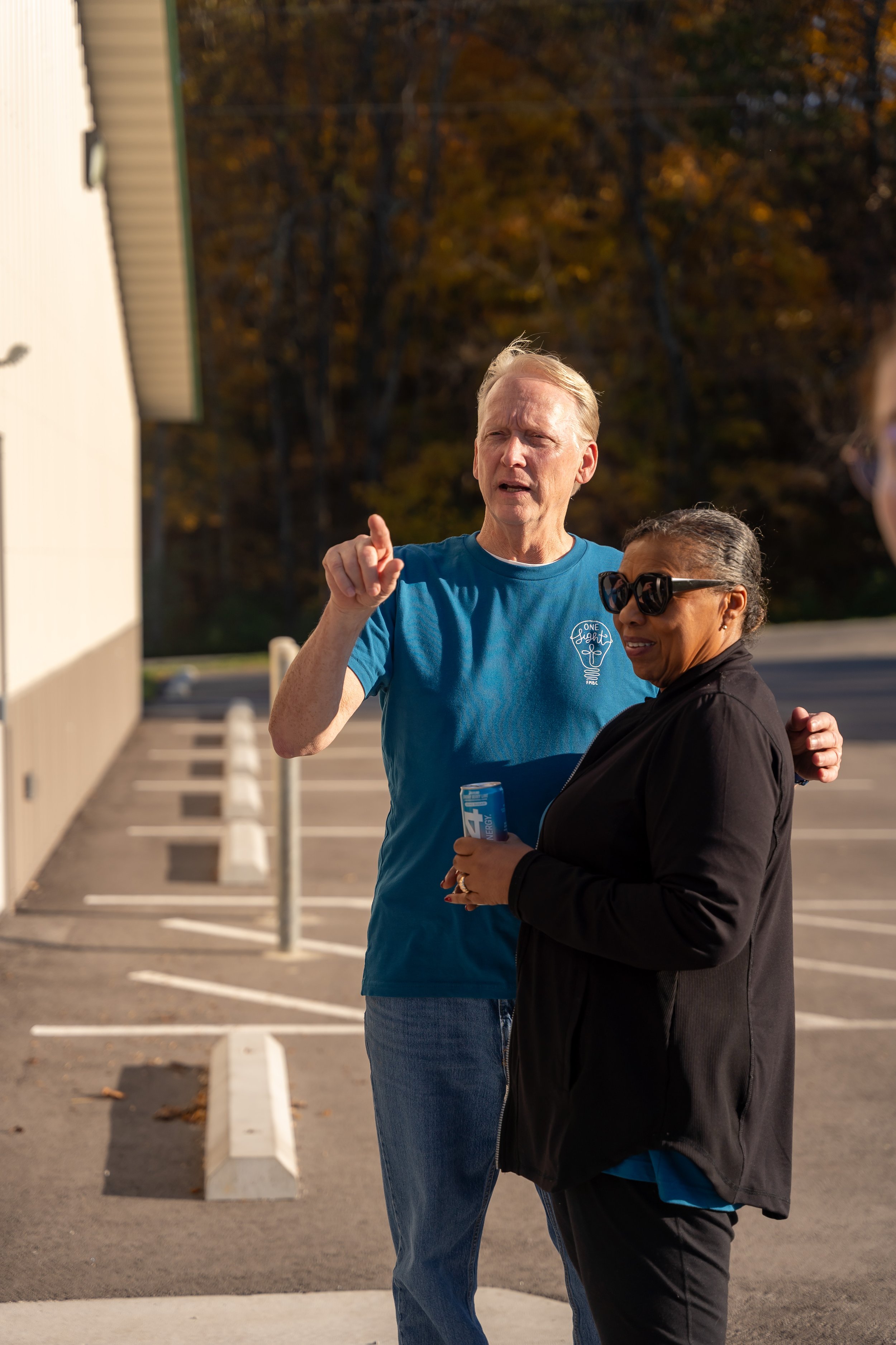 A man with blonde hair pointing and talking to a woman with dark hair wearing sunglasses, holding a can of energy drink, outside in a parking lot with striped parking spaces and barrier posts, during sunset with trees in the background.