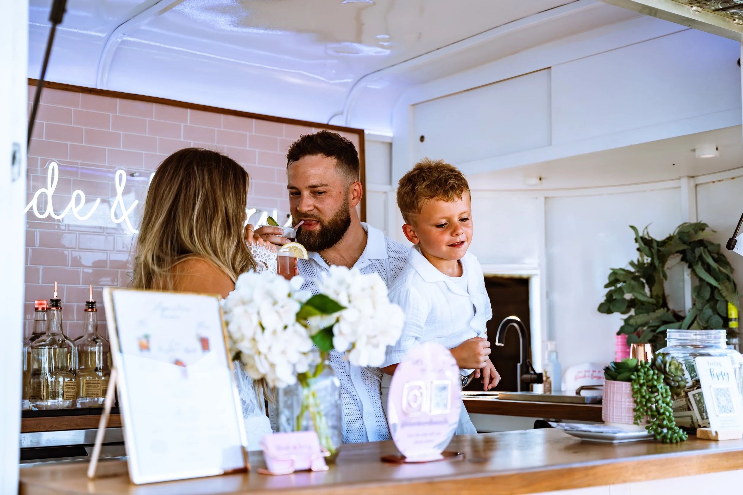 A family at a decorated dessert stand inside a bright, modern space with pink brick walls, neon sign, and lush green plants.
