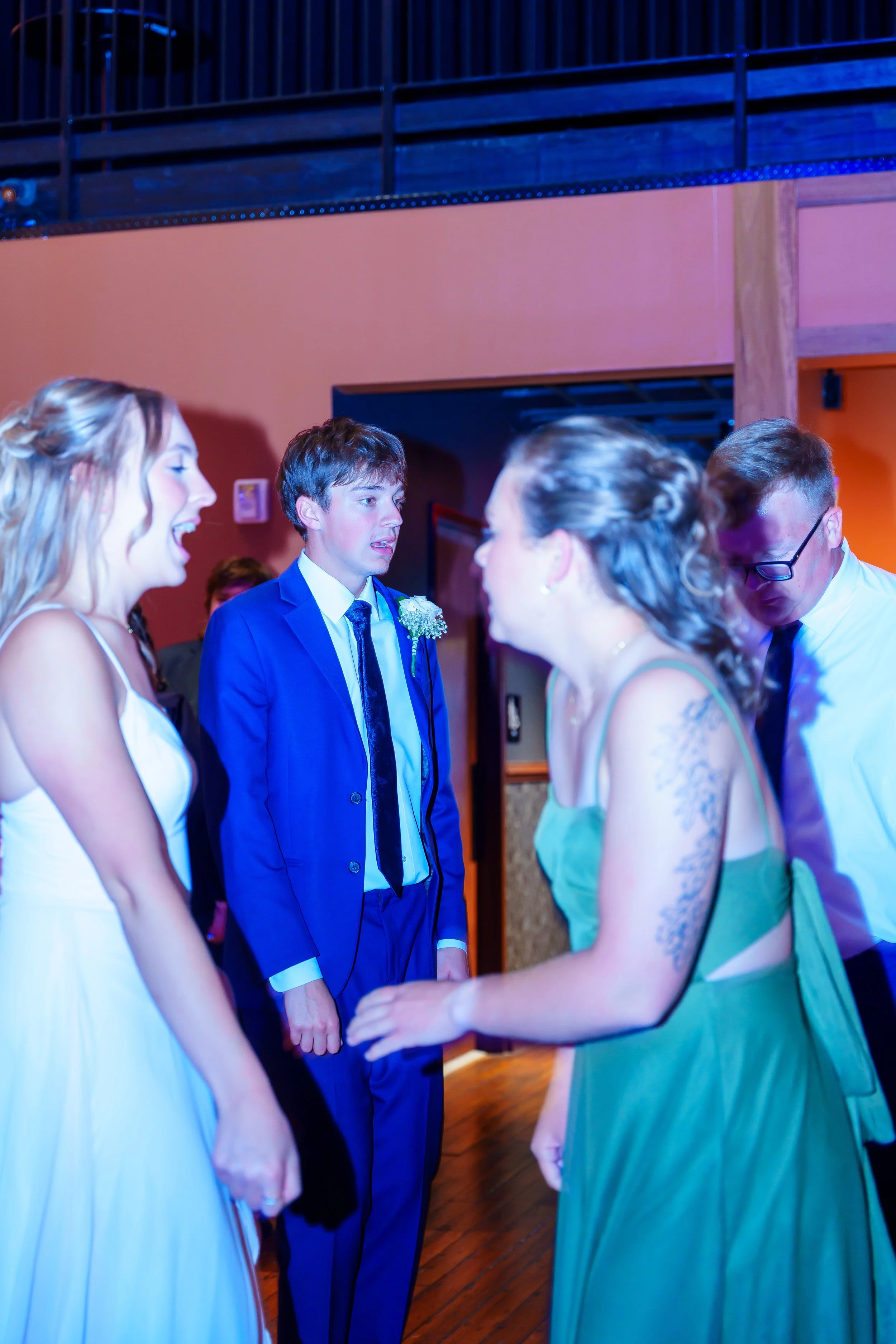 People dressed in formal attire, holding hands, engaging in a dance or ceremony indoors with colorful lighting.